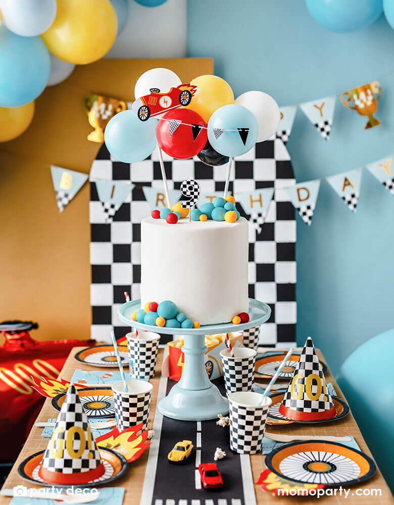 A kid's race car themed party table filled with race car themed party supplies from Momo Party including wheel shaped plates, checkered flag GO! party hats, blue race car large napkin, and checkered party cups. On the table there is a modern white birthday cake topped with Party Deco's race car balloon topper. In the back you can see a checkered backdrop with Momo Party's race car trophy happy birthday banner along with some colorful balloon garland hung on it, perfect for kid's Two Fast or Fast One party.