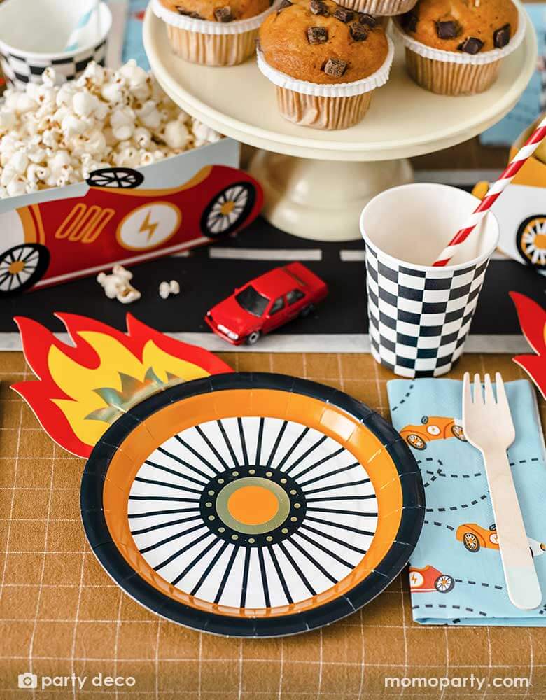 A kid's race car themed party table filled with race car themed party supplies from Momo Party including wheel shaped plates, pastel blue race car large napkin, and checkered party cups. On the table there are muffins topped with race car themed cupcake toppers and a red vintage race car shaped snack holder filled with popcorn. All laid on a brown checkered tablecloth adorn with race car track in the middle with some toy race cars, making these a perfect inspo for a rad kid's race car birthday party!