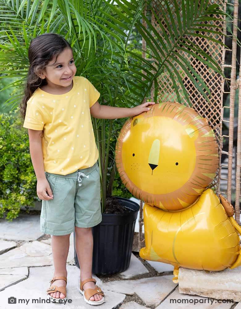 A little girl dressed in her safari adventure themed outfit standing next to Momo Party's 24" lion shaped foil mylar balloon. This charming lion shaped balloon adds the perfect amount of wild to a birthday celebration that will have your guests exclaiming "Oh My!"