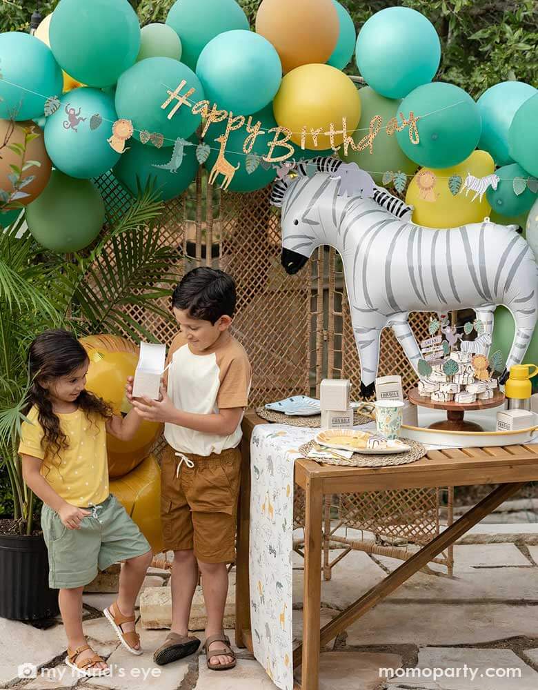 A festive kid's safari themed birthday party set up feature Momo Party's 24" zebra shaped foil mylar balloon by My Mind's Eye. Above it is a safari themed balloon garland in green, orange and yellow adorned with safari animal happy birthday banner. Next to the table which features multiple safari themed tableware and some zebra cakes in the middle are two kids in safari themed outfits opening Momo Party's cargo party favor boxes for some goodies.