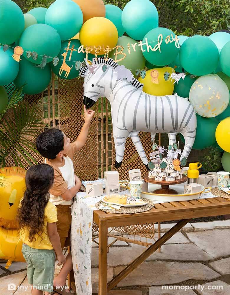 A festive kid's safari themed birthday party set up feature Momo Party's 24" zebra shaped foil mylar balloon by My Mind's Eye. Above it is a safari themed balloon garland in green, orange and yellow adorned with safari animal happy birthday banner. Next to the table which features multiple safari themed tableware and some zebra cakes in the middle are two kids pretending to feel the zebra and pet him.