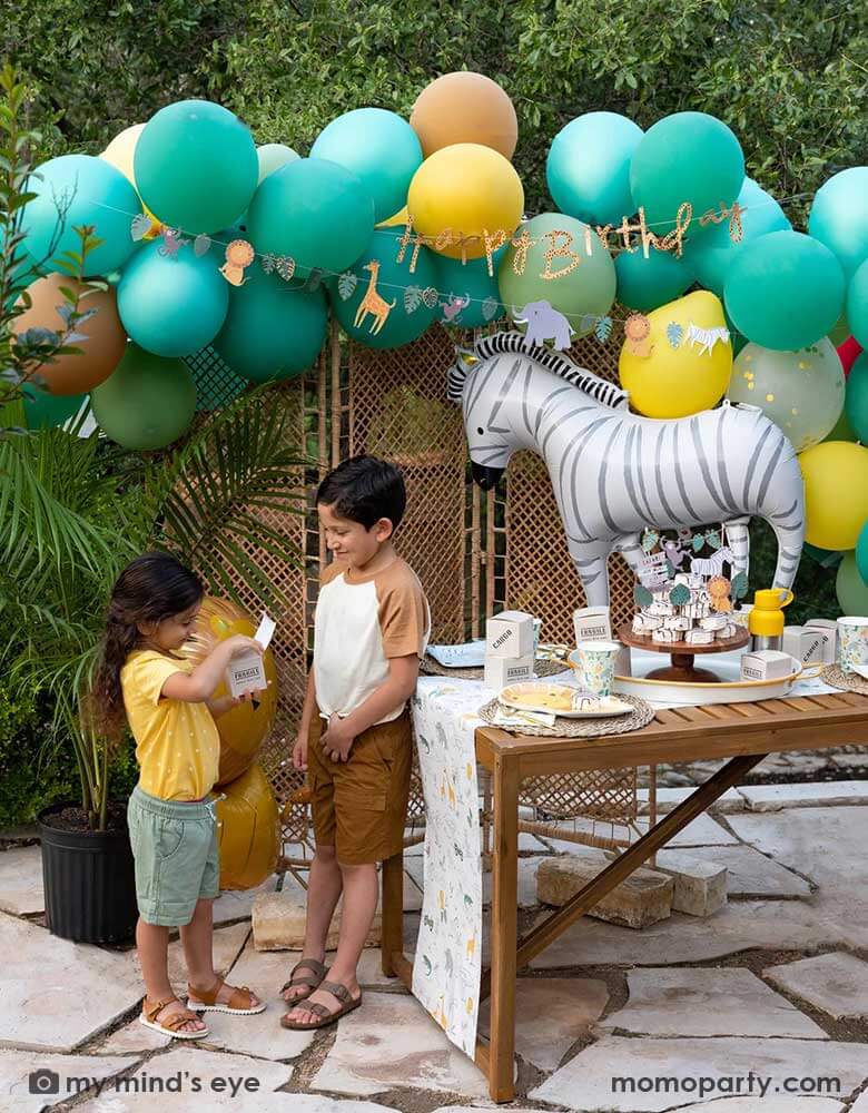 A festive kid's safari themed birthday party set up feature Momo Party's 24" zebra shaped foil mylar balloon by My Mind's Eye. Above it is a safari themed balloon garland in green, orange and yellow adorned with safari animal happy birthday banner. Next to the table which features multiple safari themed tableware and some zebra cakes in the middle are two kids in safari themed outfits opening Momo Party's cargo party favor boxes for some goodies.