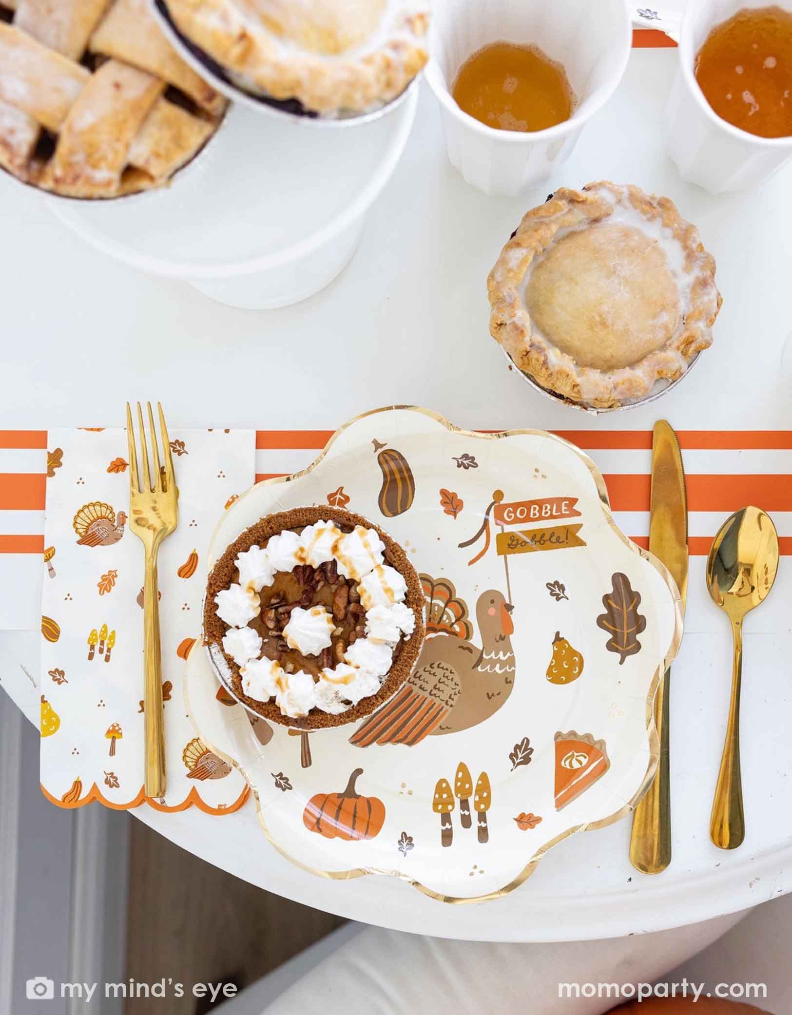 A Thanksgiving-themed party table featuring various Thanksgiving themed party supplies from Momo Party including turkey plates, napkins, gold cutlery, orange striped table runner, party cups with fall themed designs on them. With decorative elements and fall themed desserts and pumpkin pies on a table.