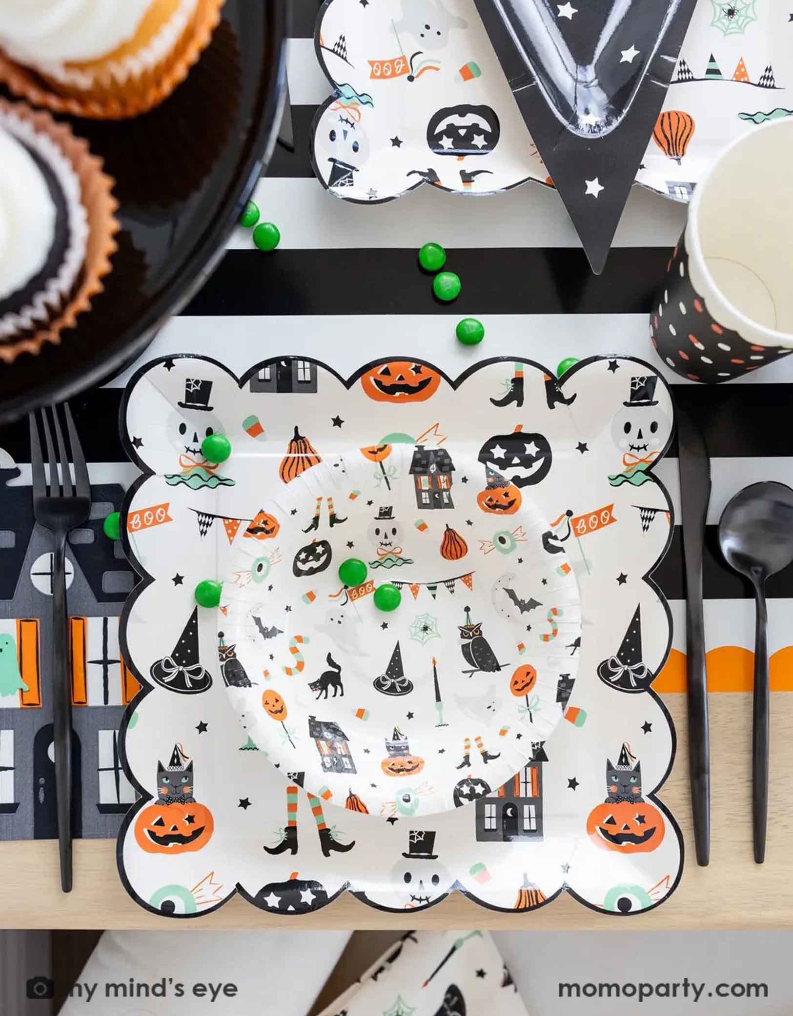 A overhead shot of a festive Halloween-themed table setting with Momo Party's Trick or Treat dinner plates and bowls, the haunted mansion shaped napkins and black cutlery set on a black orange and white striped table runner fro Momo Party. On the table there's a cauldron filled with green gum balls and some green candies spread across the table. A perfect set up for kid's friendly Halloween celebration.