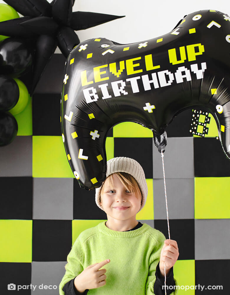 A young boy in lime green colored sweater with black long sleeves holding Momo Party's 25.5 "x 18" video controller shaped foil balloon featuring LEVEL UP BIRTHDAY message. Behind him is a video game themed party backdrop with black, grey and lime squares adorned by a balloon garland in black and lime colors and a jumbo black starburst foil balloon - a perfect decoration for a kid's video game themed birthday party.