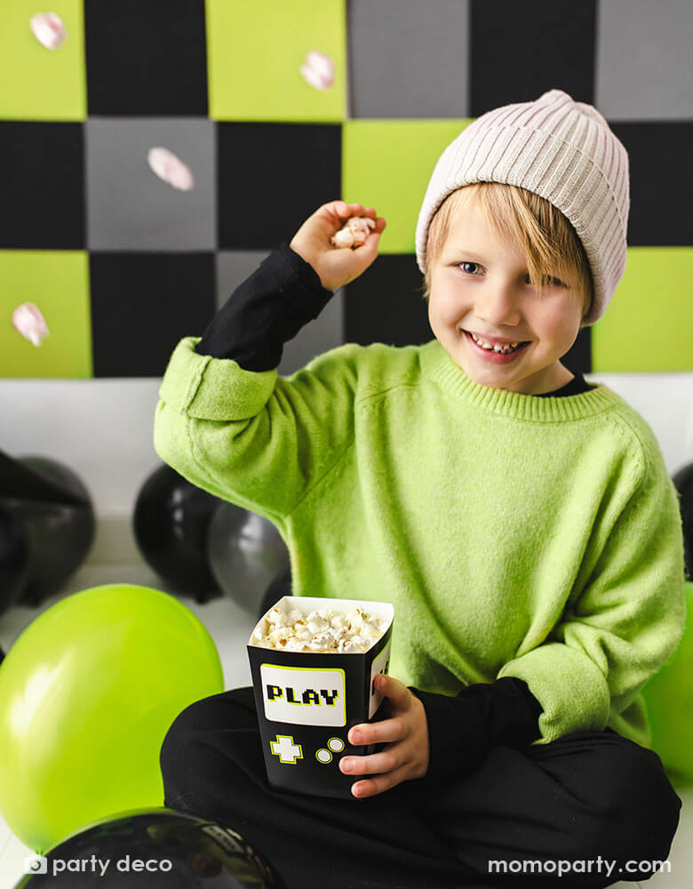 A boy in the classic video game black and green outfit sitting on the floor surrounded by black and green party balloons. He's holding Momo Party's 2.75 x 2.75 x 4.75 inches black video game snack box by Party Deco, filled with popcorn, trying to throw the popcorn at his friend.