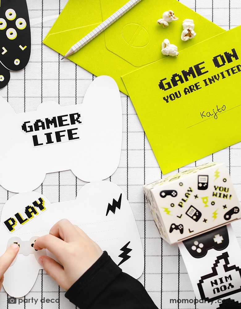 A boy making a video game themed birthday invitation using Momo Party's Gamer Sticker Roll by Party Deco. Each roll includes 36 stickers in 18 design including video game devices and game controllers. These stickers are perfect for party favors and activities at any gaming party.