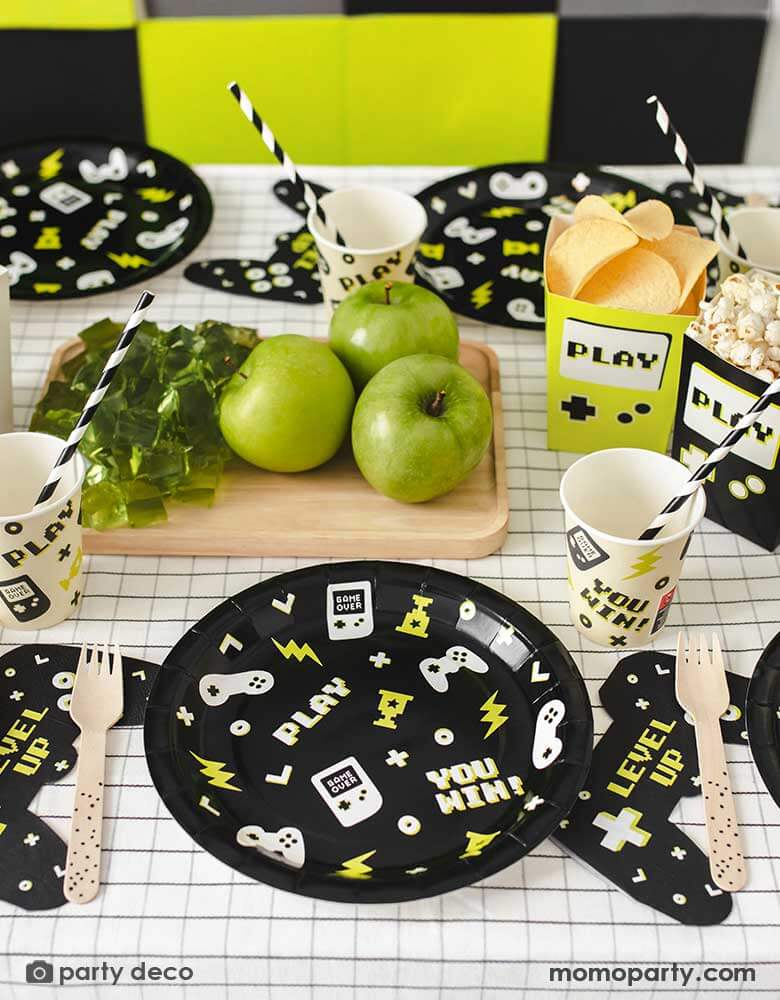 A video game themed party table featuring Momo Party's black Video Game round party plates with video game devices and controller motifs on them. Next to the plates are a game controller shaped napkins with "LEVEL UP" message on them, with the coordinating video game party cups with black striped paper straws from Momo Party. In the middle of the table, there are some green apples and green jellos next to two game boy shaped paper snack box filled with popcorn and chips.