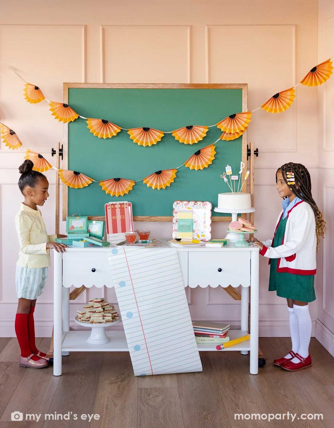 Two girls standing in front of a school themed decorated party table with a cake, surrounded by various school themed party supplies and decorations from Momo Party including party plates, napkins, table runner in front of a chalkboard above which is decorated with pencil party fans garland hung above, making this perfect inspiration for kid's first day of school celebration or back to school party.