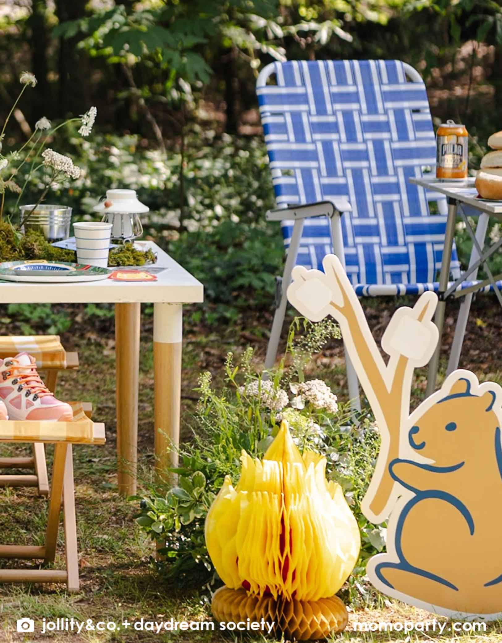 A kid's camping themed party in the outdoor setting with a kid's camping table, chair, and decorative camping themed decorations from Momo Party including camping inspired tablewares like plates, napkins and party cups and camp fire honeycomb decoration and a squirrel shaped cutout holding marshmallows - a whimsy and adorable setting that's great for kid's birthday celebration. 