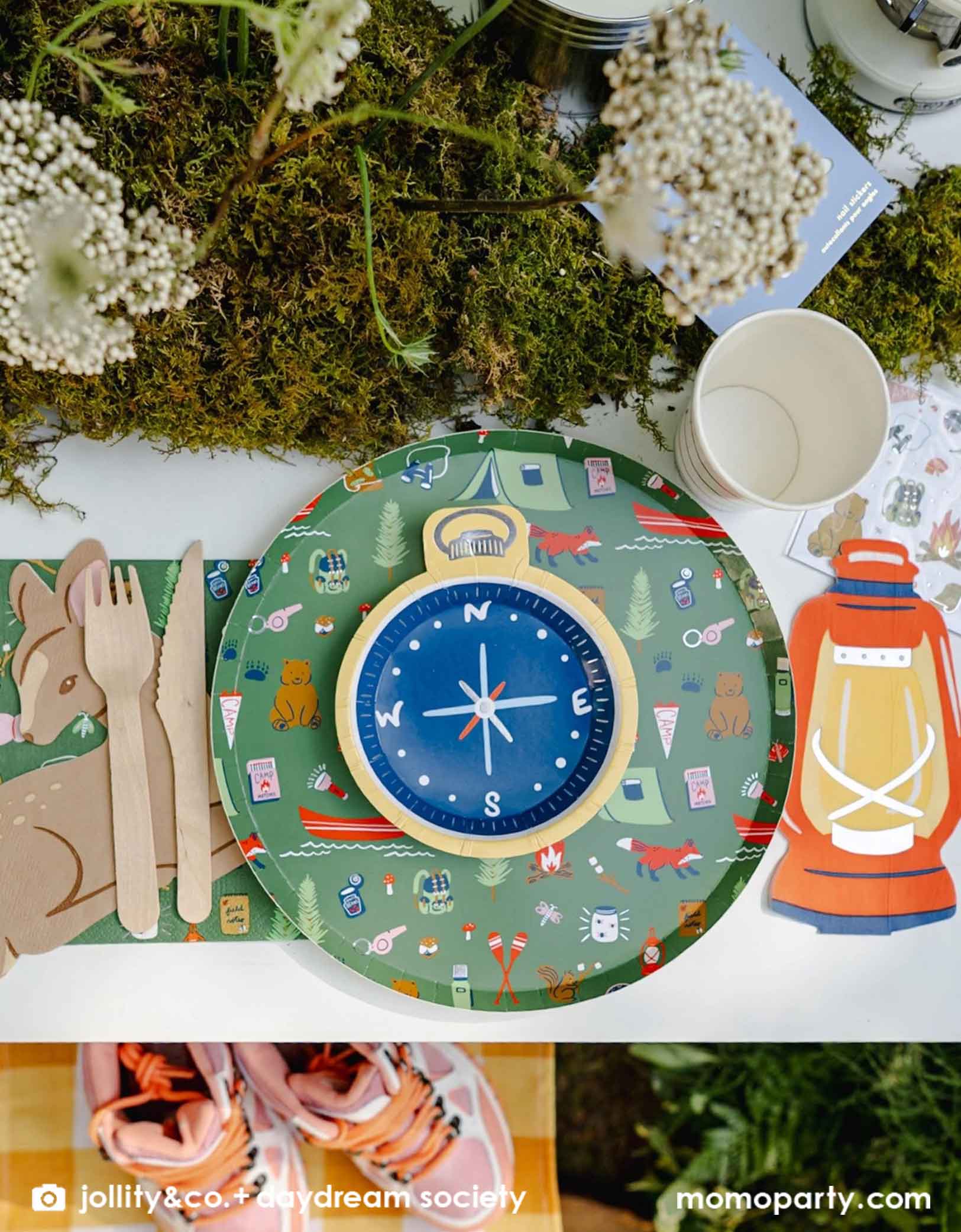 A children's camping themed party table setting with Momo Party's compass shaped mini plate, camping themed patterned dinner plates and napkins, camping lantern shaped guest napkins, deer shaped napkins, wooden cutlery, and paper cups on a moss-decorative background.
