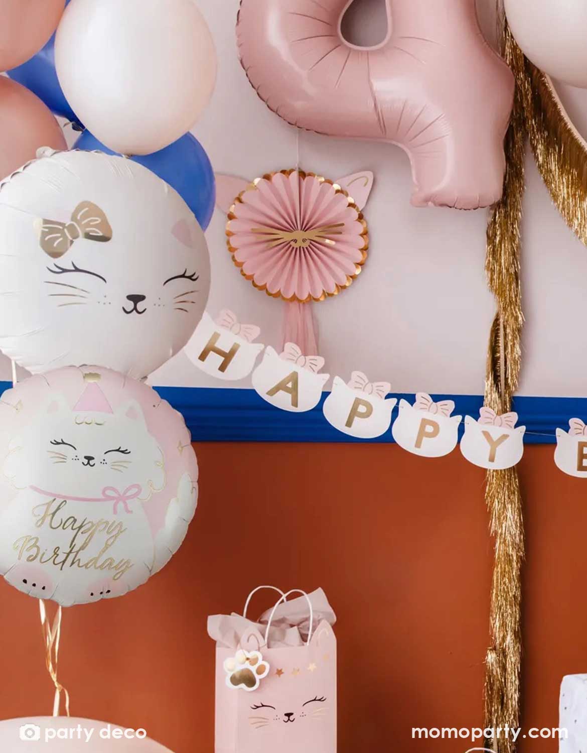 A kid's cat themed birthday party decoration set up features Momo Party's cat themed party supplies by Party Deco including two 18" round cat foil balloons in light pink, along with a latex balloon garland in pink, blush and royal blue. On the wall there are pink kitty cat shaped party fans along with a gold tinsel garland hung on. With a pink number 4 foil balloon and a cat head shaped happy birthday banner, it makes an adorable inspo a cat or pet themed party.