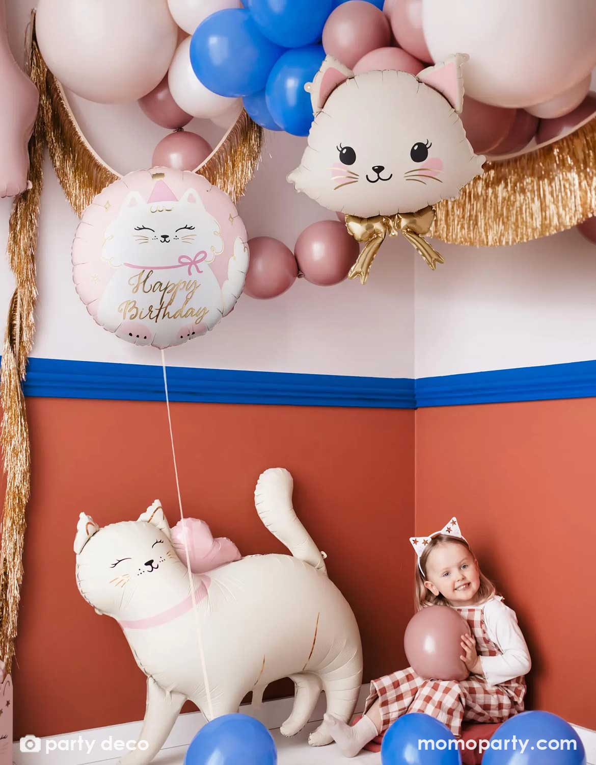 A kid's cat themed birthday party decoration set up features Momo Party's cat themed party balloons by Party Deco including a cat shaped foil balloon in light pink, along with a latex balloon garland in pink, blush and royal blue and some gold tinsel garlands. Next to it there's a pink round foil balloon with cat design and happy birthday on it. On the floor there's a girl holding a balloon with a  standing cat shaped foil balloon next to her, making this an adorable set up for a kid's cat themed party.