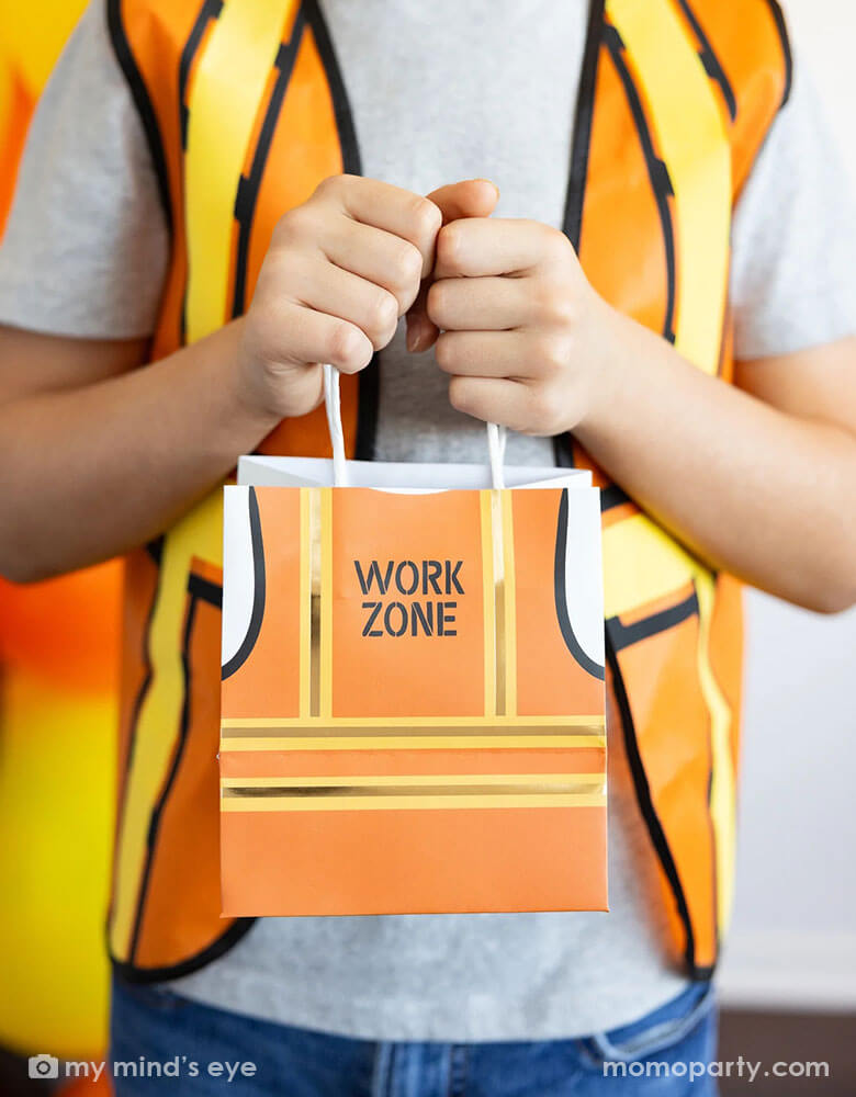 A boy dressed up as a construction worker holding Momo Party's 5.5" x 5" x 4" Construction Vest Treat Bags by My Mind's Eye. These treat bags will make any party a blast with their playful construction vest design. Perfect for construction themed parties or just for fun, this set will be a hit with kids and adults alike. 