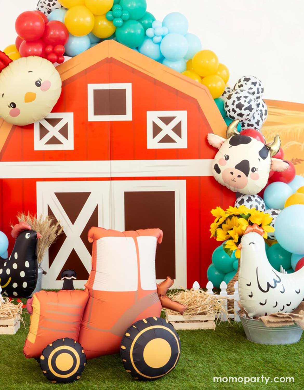 A kids farm themed party backdrop decoration featuring a red barn house backdrop adorned with colorful farm inspired balloons in festive colors and various sizes, along with farm animal shaped foil balloons including a chicken head shaped balloon, a cow shaped balloon, a hen shaped standing foil balloon , and a red tractor shaped standing balloon. 