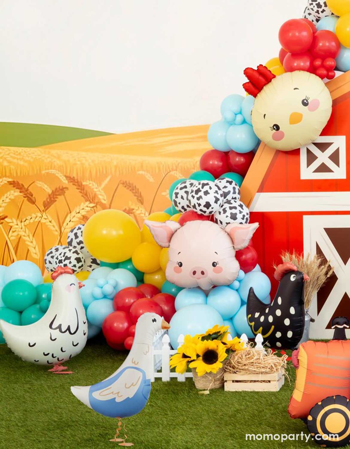 A kids farm themed party backdrop decoration featuring a red barn house backdrop adorned with colorful farm inspired balloons in festive colors and various sizes, along with farm animal shaped foil balloons including a chicken head shaped balloon, a pig head shaped balloon, a hen shaped standing foil balloon , and goose shaped standing standing balloon on a lawn.