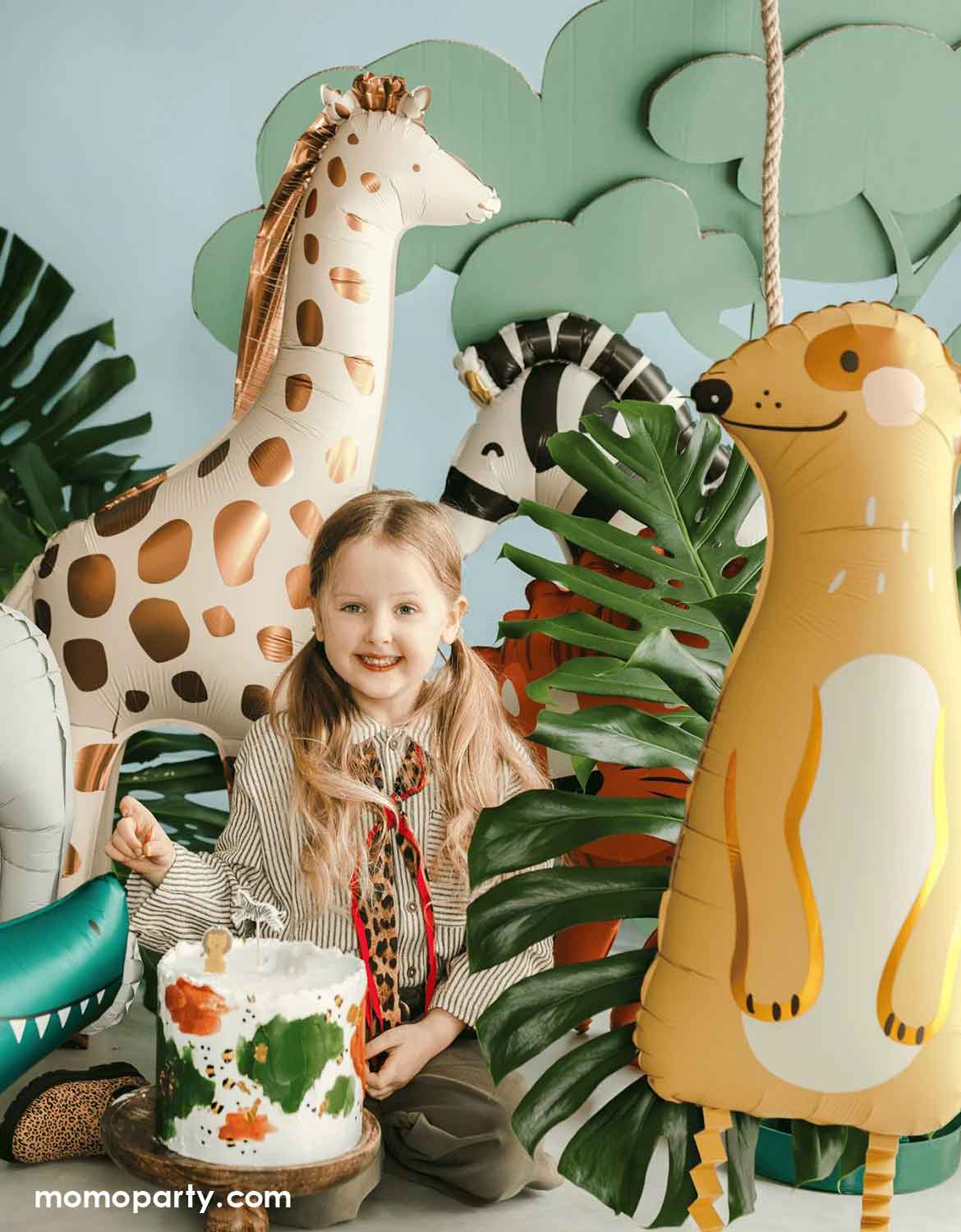 A little girl with safari adventure inspired outfits with a birthday cake surrounded by animal themed foil balloons from Momo Party including a giraffe, a zebra and a meerkat in a jungle-themed setting with palm leaves around.