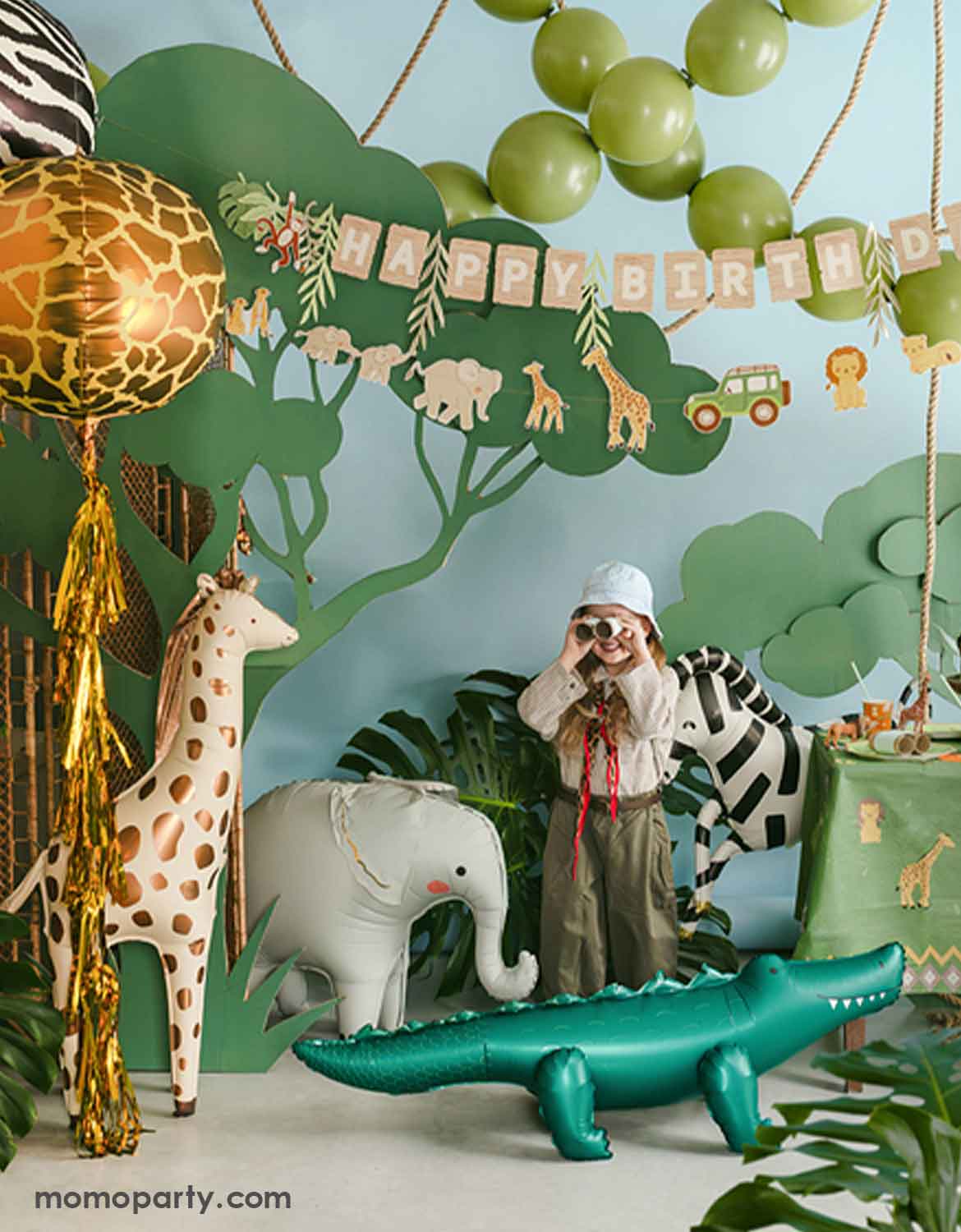 A child in safari adventure inspired outfits with binoculars at a safari jungle themed birthday party setting featuring  animal themed foil balloons from Momo Party including a standing giraffe, a zebra, an elephant, an alligator, and a couple of orb shaped animal print round balloons with gold fringe attached in a jungle-themed setting with palm leaves around. In the back of the wall, there's a safari themed happy birthday banner hung above along with a green balloon garland.