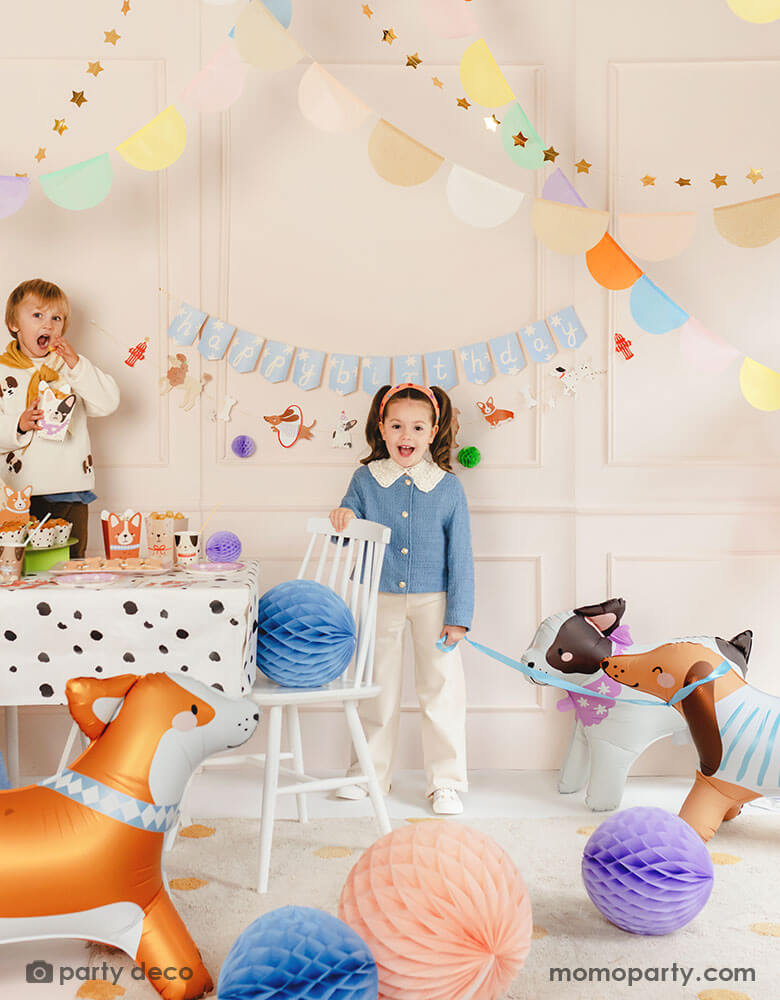 A kid's puppy dog themed birthday party set up featuring various dog themed party supplies from Momo Party including party banners, paw balloons, corgi, bull dog and Dachshund standing foil balloons, Dalmatian print dots tablecloth, dog shaped snack boxes, party cups and honeycomb decorations in soft pastel colors. These cute dog themed party goods make a festive touch to a kid's birthday bash!