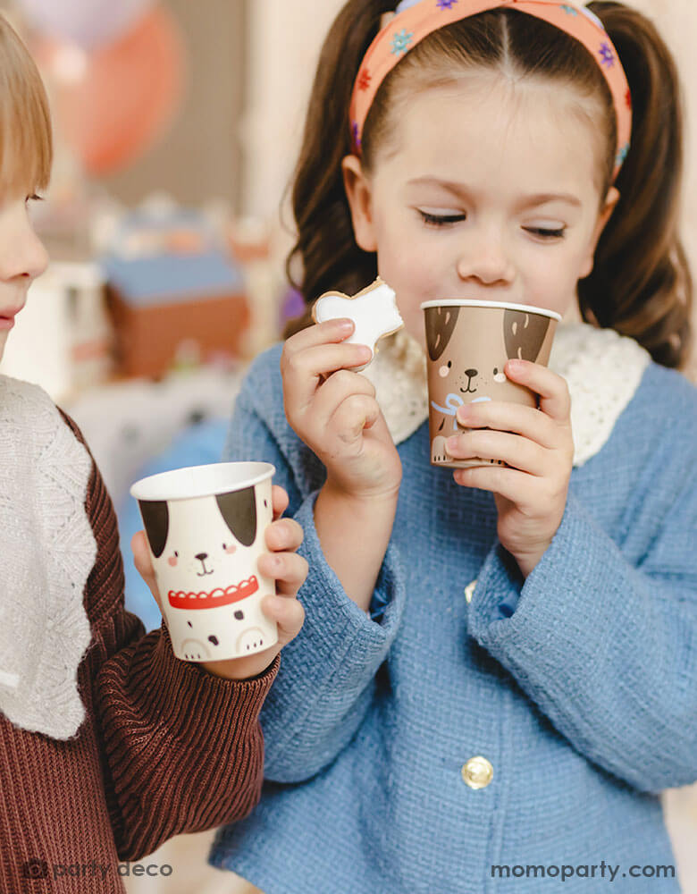 Two preschooler kids in a puppy dog themed birthday party holding Momo Party's 7.4 oz dog party cups with adorable puppy design on them, sipping their drinks and having fun. The girl is also holding a bone shaped sugar cookie that goes with her beverage. 