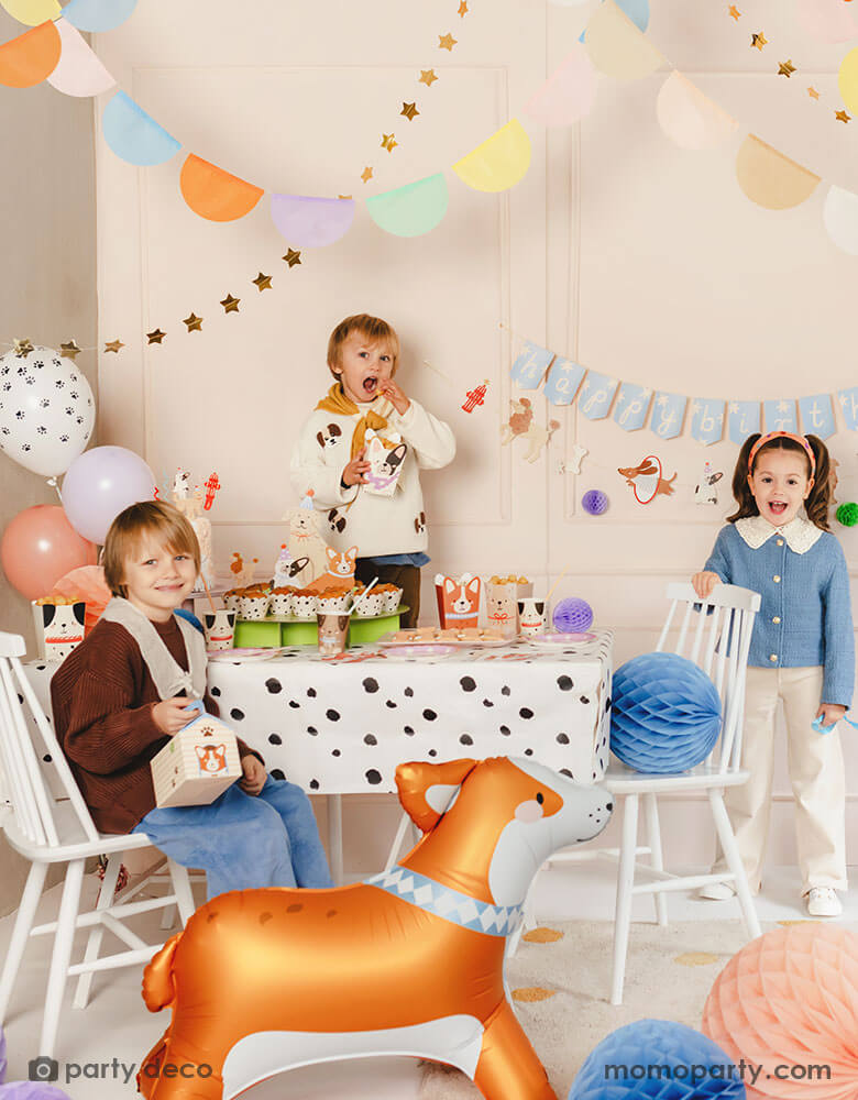 A kid's puppy dog themed birthday party set up featuring various dog themed party supplies from Momo Party including party banners, paw balloons, corgi standing foil balloon, Dalmatian print dots tablecloth, puppy dog shaped snack boxes, party cups and honeycomb decorations in soft pastel colors. These cute dog themed party goods make a festive touch to kid's birthday bash!