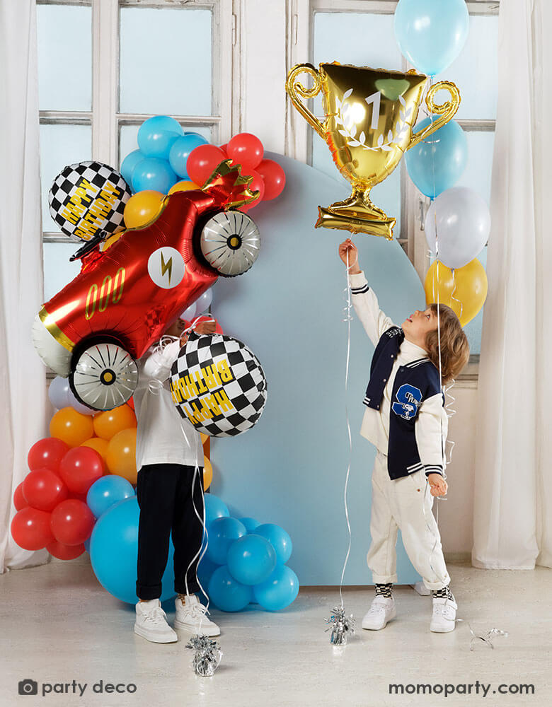 Two boys playing with Momo Party's race car themed foil balloons including a red vintage race car shaped balloon, two checkered flag happy birthday foil balloons and a gold trophy shaped foil balloon, both kids standing in front of a pastel blue party backdrop wall which is decorated with colorful balloon garland with race car inspired colors including red, light blue, orange, gray and yellow. All makes this a festive and fun kid's birthday celebration.