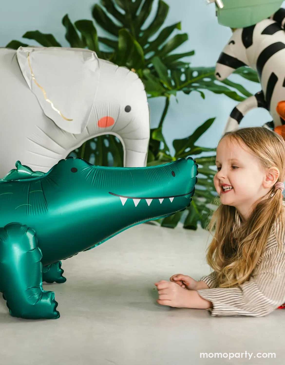 A young girl with blonde hair smiles while looking at a shiny teal inflatable alligator balloon. In the background, other animal balloons, including a white elephant and a zebra, are visible against a backdrop of green leaves and a light blue wall.