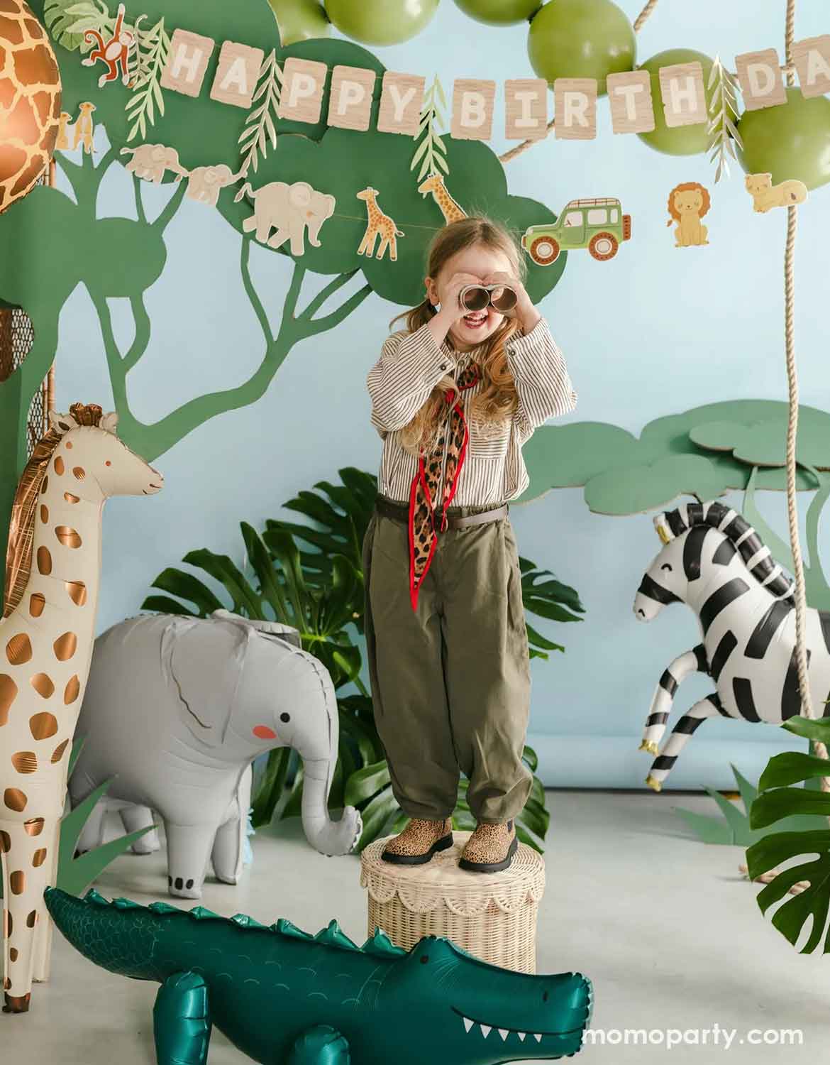 A young girl stands on a woven stool in a jungle-themed children's party setting, wearing safari attire with a cream knit top, olive green pants, and a red patterned neck scarf, holding toy binoculars. Around her are multiple safari animal foil balloons from Momo Party including a giraffe, an elephant, a zebra and a alligator, along with happy birthday party banner hung above.
