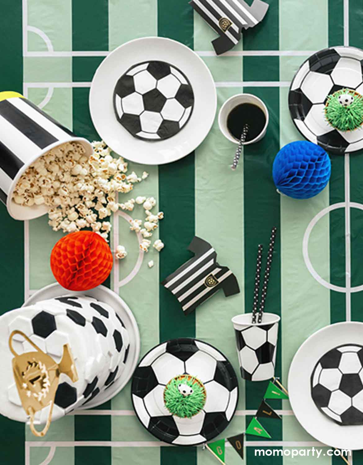 Soccer-themed party table setting with soccer shaped plates, cups, and decorations on soccer field tablecloth from Momo Party.