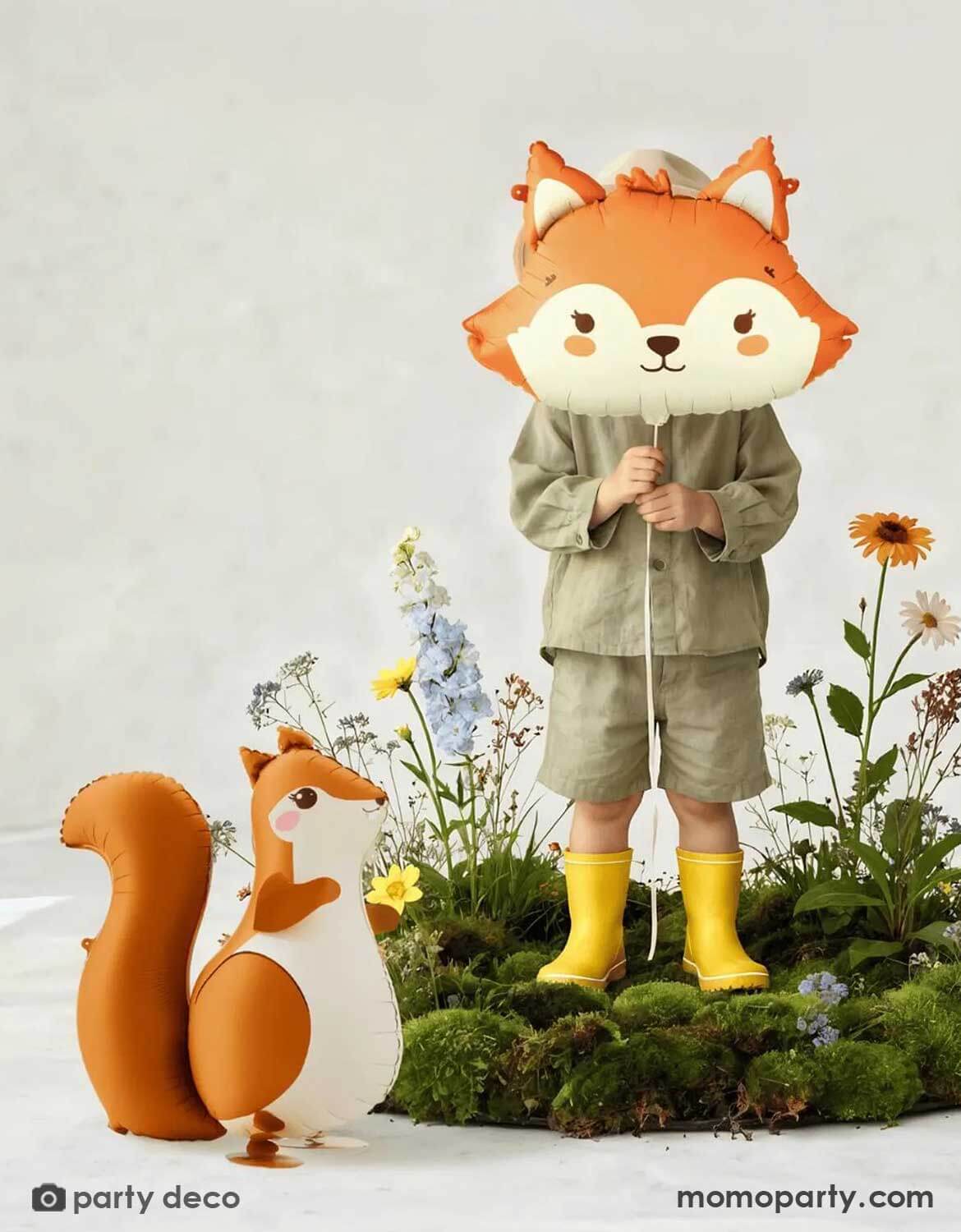 A child holding Momo Party's fox-shaped foil balloon with a decorative woodland setup featuring flowers moss. Next to it is Momo Party's 30" squirrel shaped standing foil balloon.