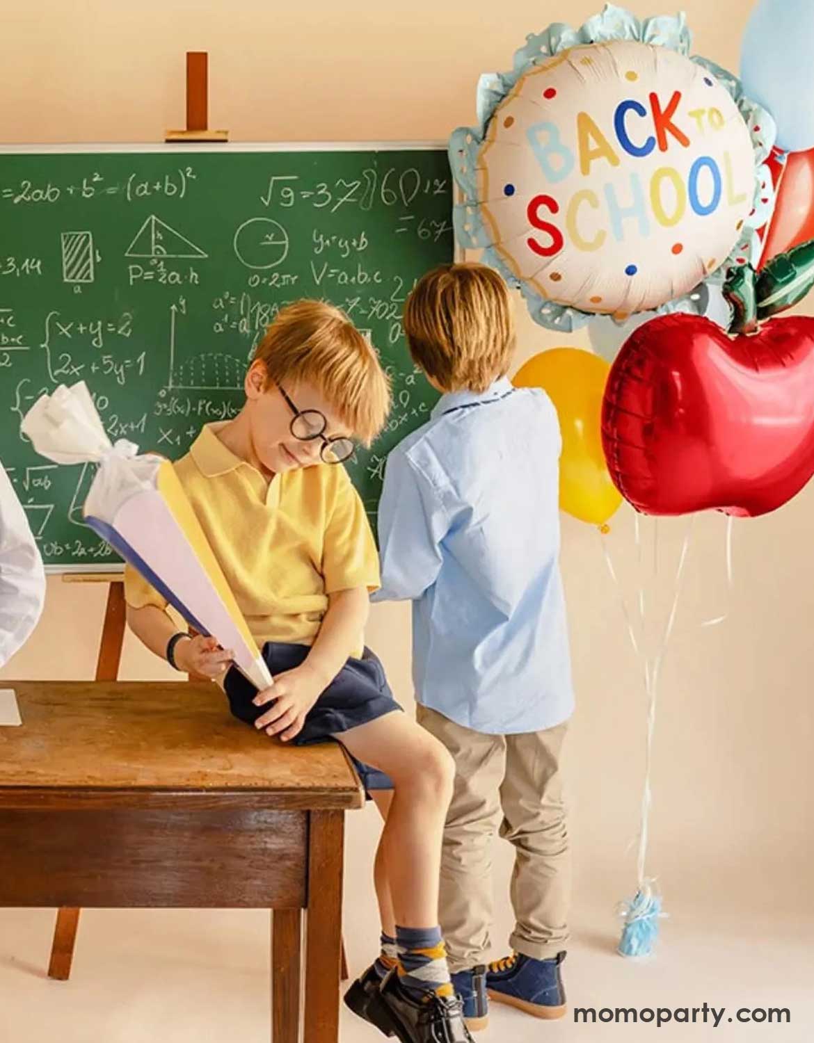 Two kids in their uniforms sitting/standing in a classroom setting with Momo Party's 20" scalloped "Back to School" foil balloon, an apple shaped foil balloon along with some festive latex balloon bundles next to a chalkboard - a perfect setup for kids back to school party, classroom decorations or first day of school celebrations.