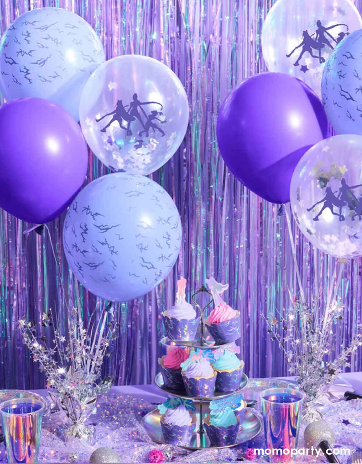 A kpop themed birthday party decoration set up featuring Momo Party's Kpop Party Latex Balloon Mix in purple, lilac and clear with silver stars and Kpop Demon Hunters figures designs against a purple foil fringe curtain backdrop, on the party table are some buttercream cupcakes wrapped in glitter purple wrappers with iridescent party cups, plates, napkins and glitter decorations around.