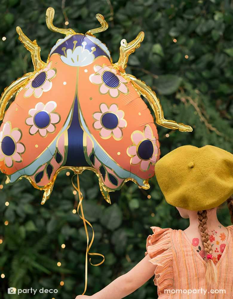 A little girl holding Momo Party's 34 x 30 inches ladybird shaped foil balloon by Party Deco. With coral color with small flower pattern with gold legs on it. She's dressed in her spring outfit with a mustard beret hat standing in a backyard with a wall filled of greenery.