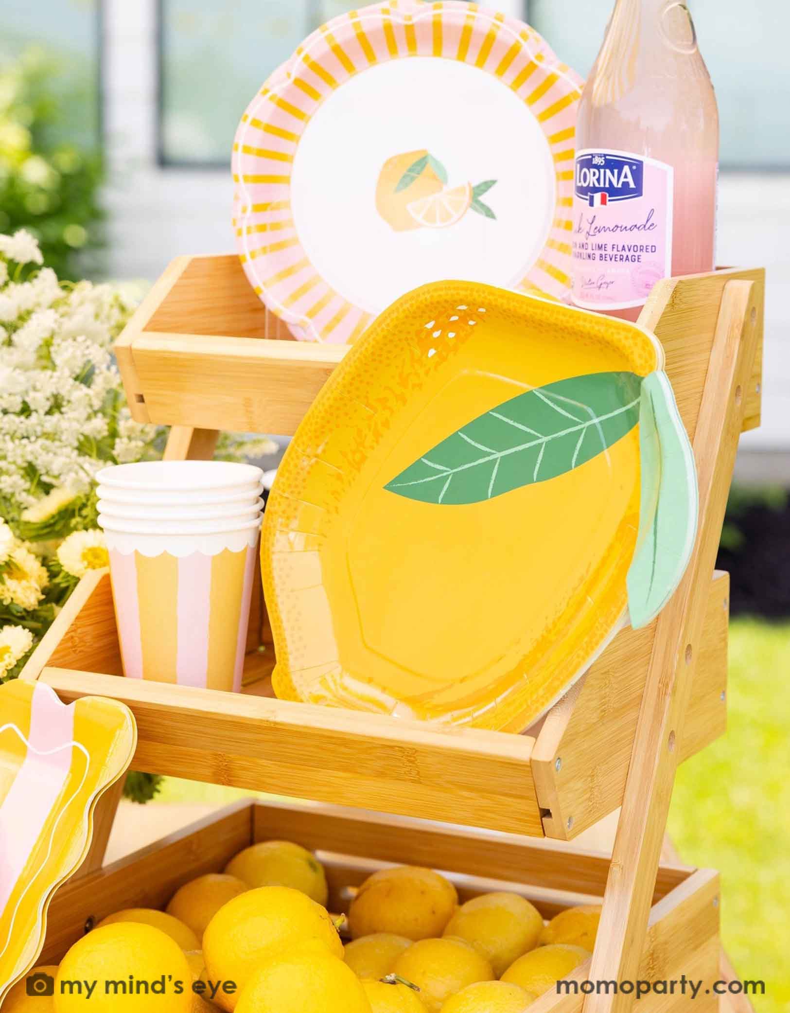 A 8.75" x 11.25" yellow lemon shaped plate with lemon themed party cups and side plates form Momo Party on a wooden stand. With a bottle of Lornia lemonade,  lemons and flowers in the background.