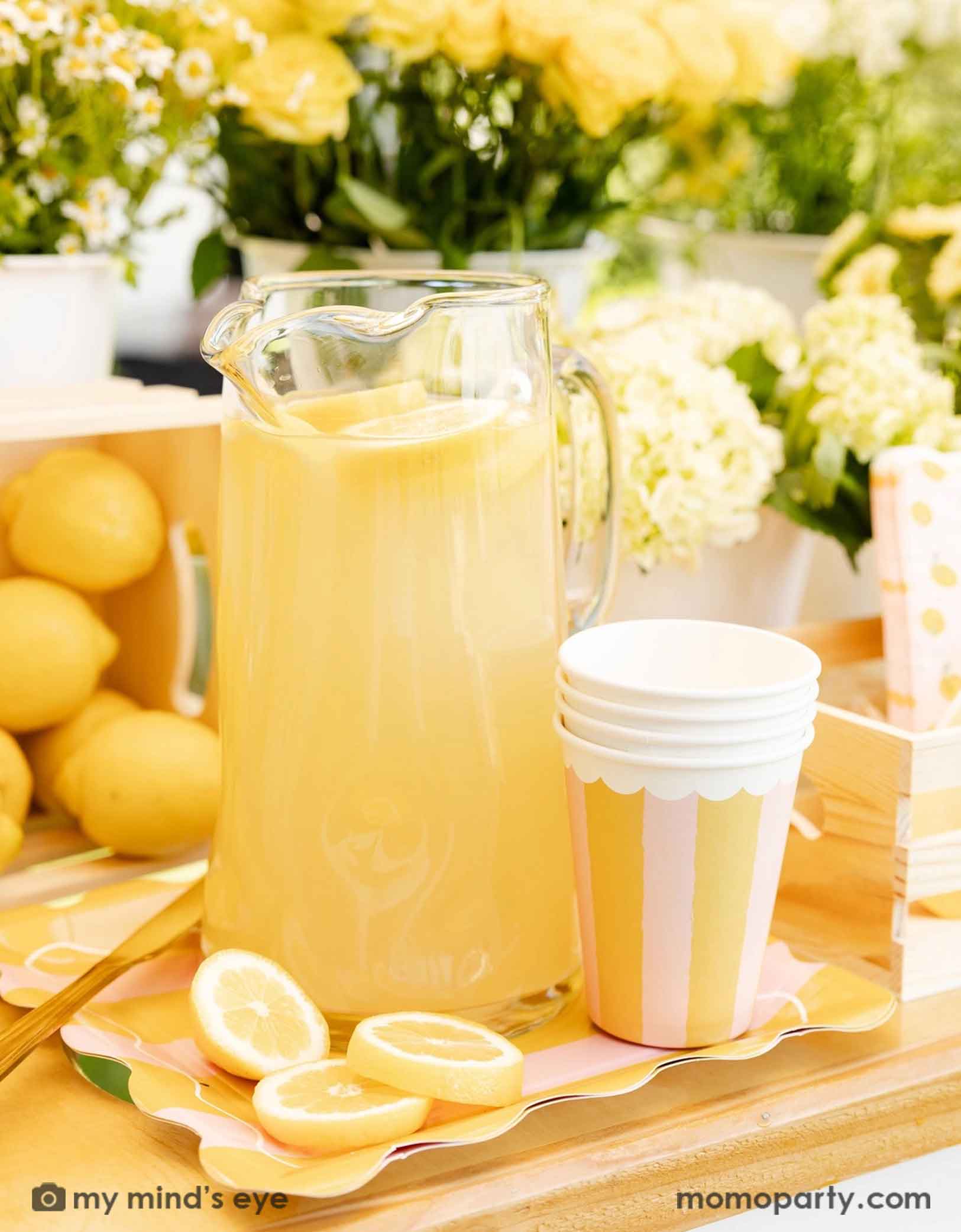 A glass pitcher of lemonade with lemons and Momo Party's Yellow & Pink Striped Party Cups next to it, on a wooden dessert cart with lemons and flowers in the background.