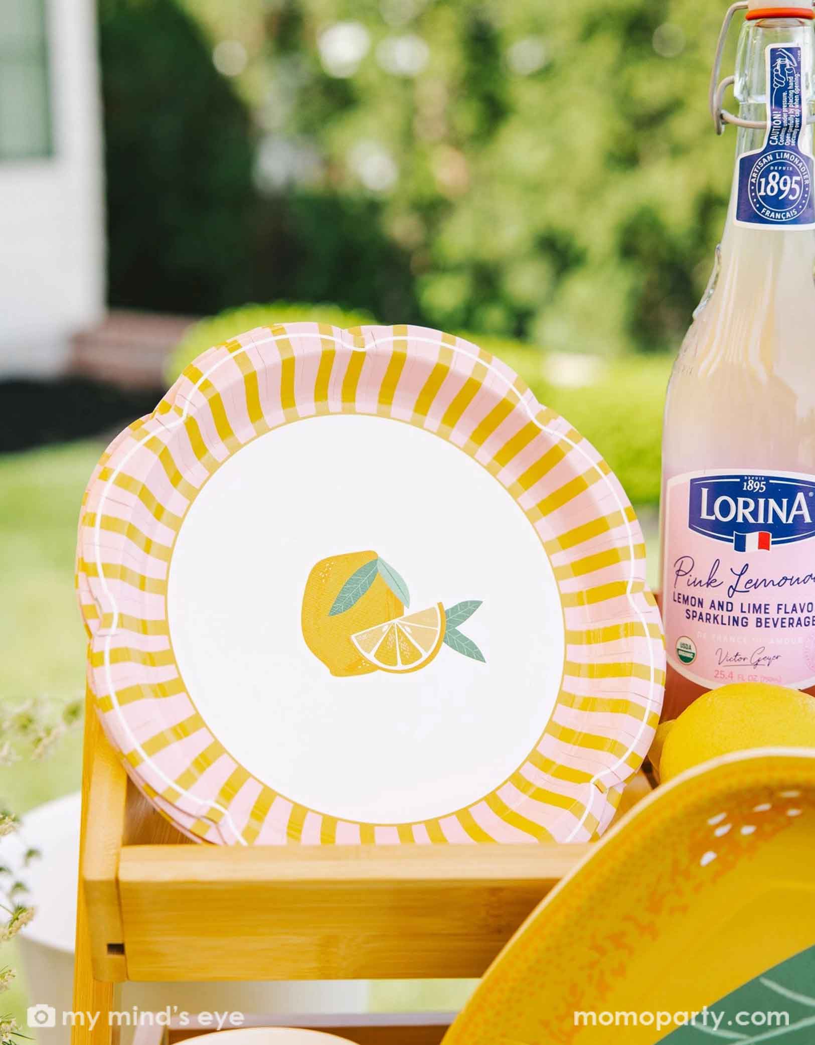 Momo Party's 9" Yellow & Pink Striped Scallops paper plate with lemon design and yellow and pink striped border by My Mind's Eye and a bottle of Lorina lemonade on a blurred outdoor background.