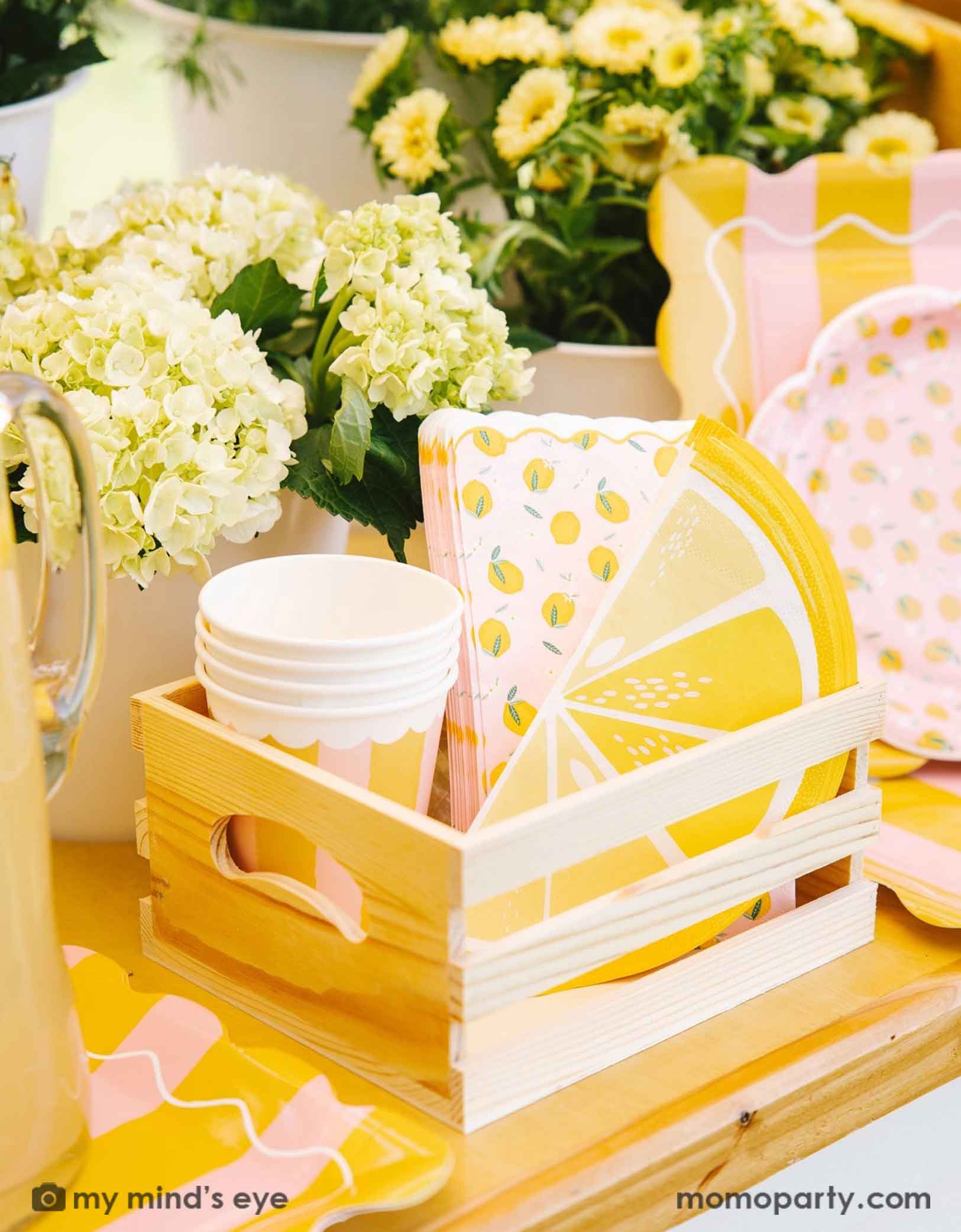 A cute wooden crate filled with party supplies with lemon design on plates, party cups and napkins from Momo Party, set against a beautiful floral background.