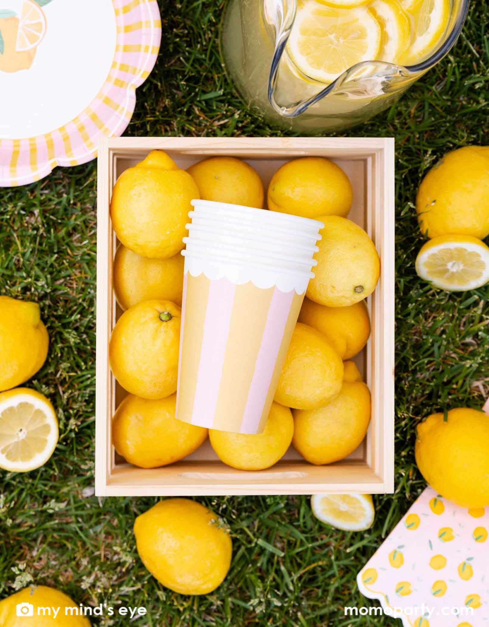 A wooden crate box of many lemons with Momo Party's Yellow & Pink Striped Party Cups in it, along with a pitcher of lemonade and lemon themed party plates and napkins on grass.