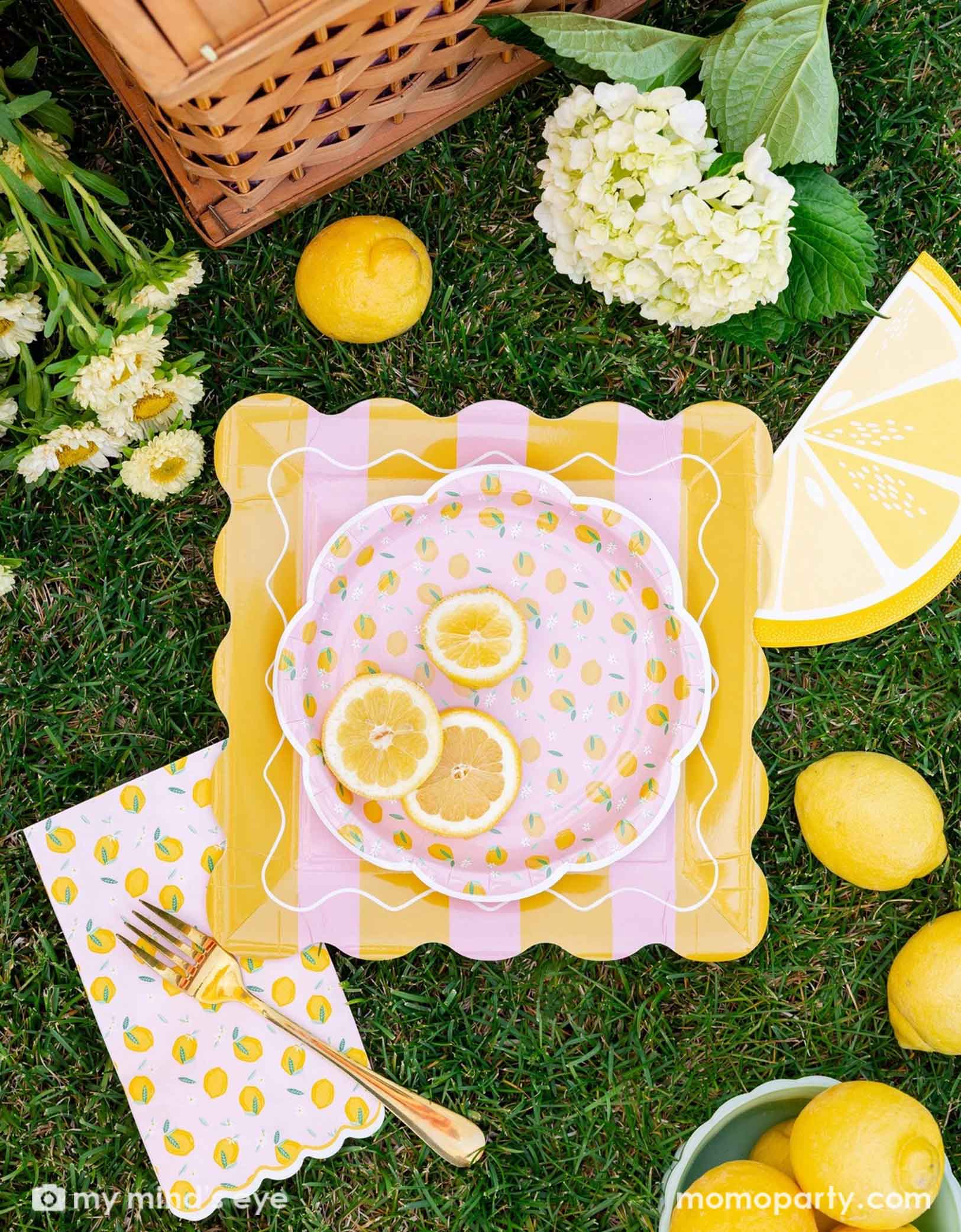 Decorative table setting featuring various lemon themed party tablewares from Momo Party including the 10" yellow and pink striped plate, lemon scattered round plate, lemon scattered dinner napkin and lemon slice shaped napkin. With flowers and lemons on a grassy surface.
