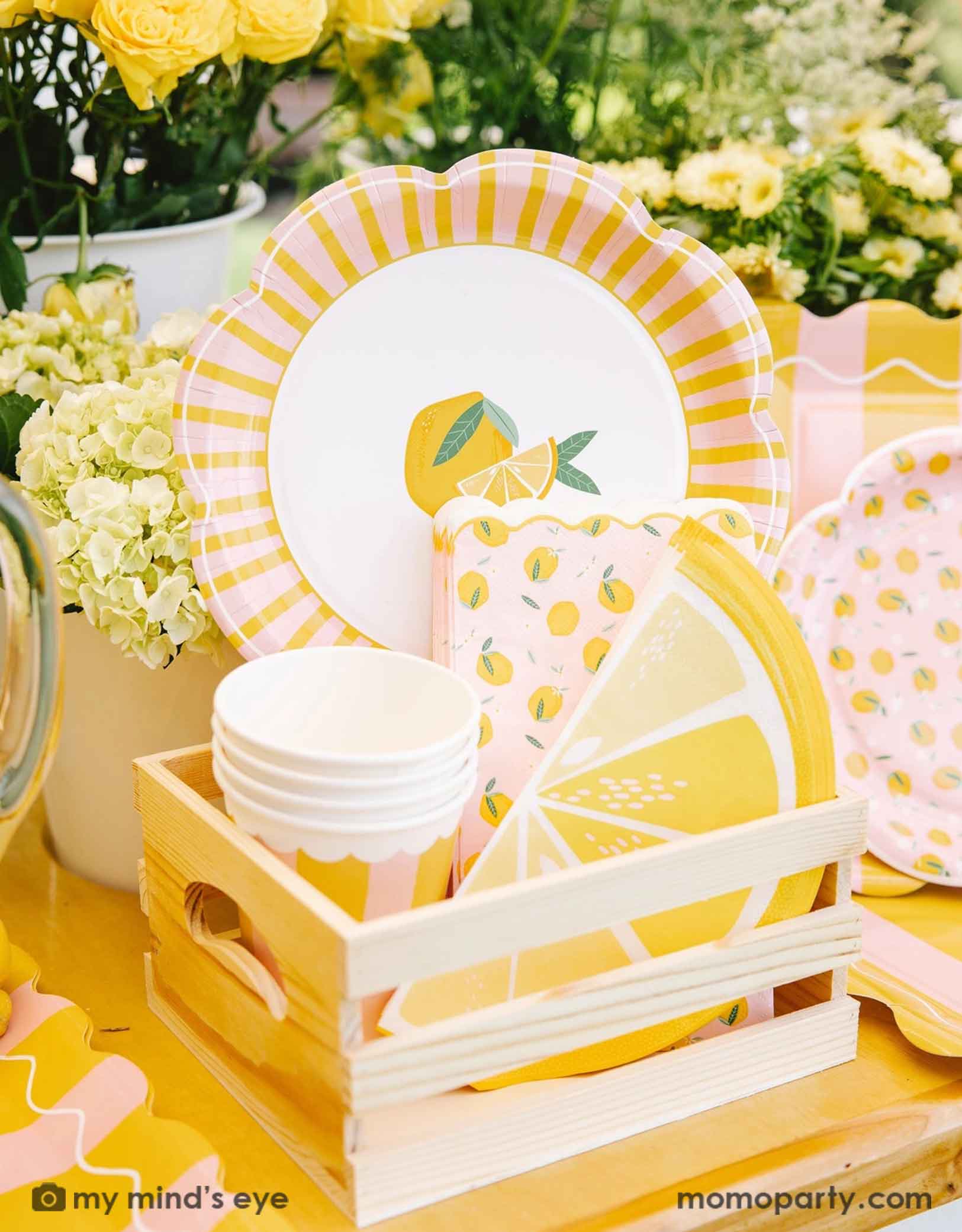 A cute wooden crate filled with party supplies with lemon design on plates, party cups and lemon sliced shaped napkins from Momo Party, set against a beautiful floral background.