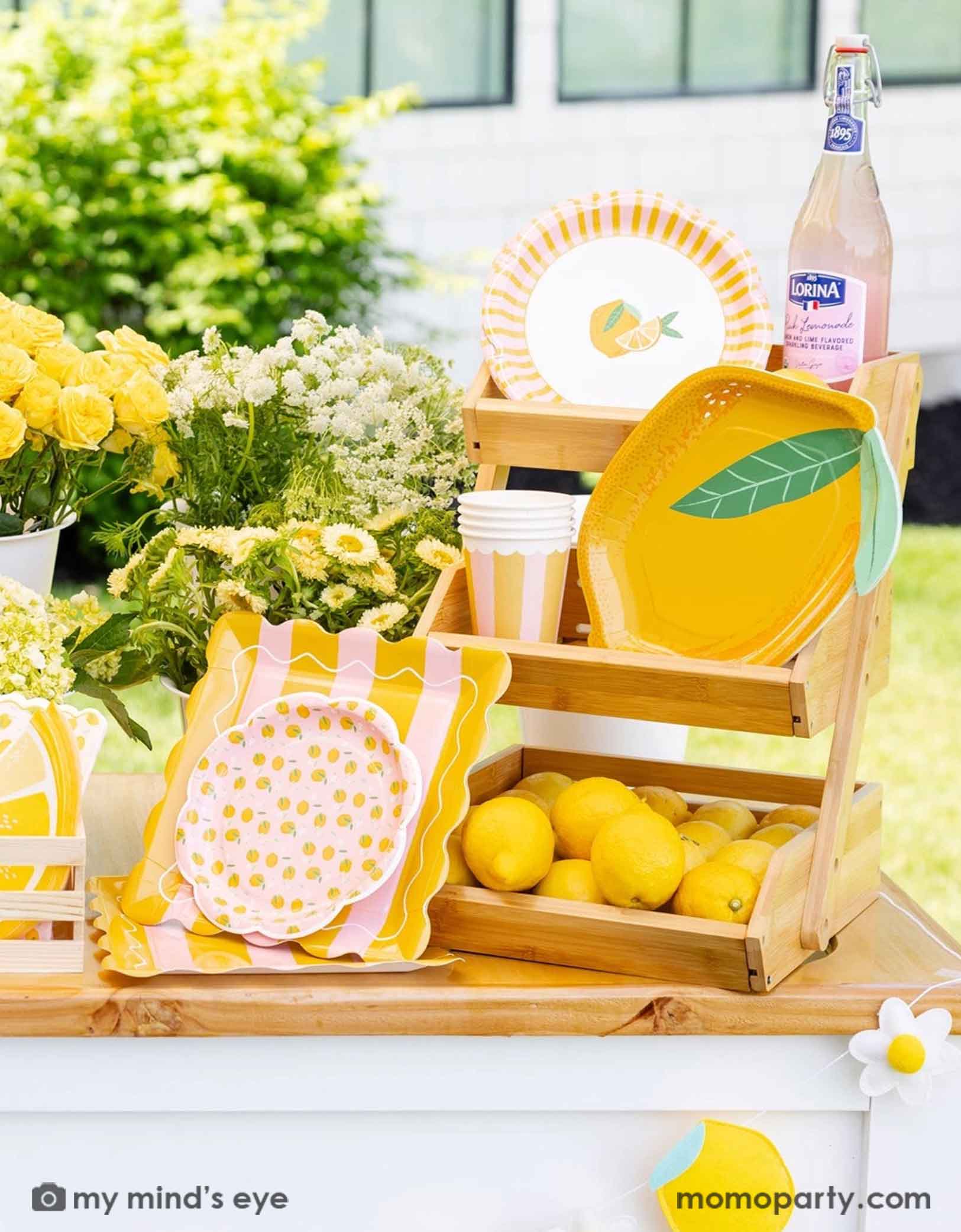 Decorative lemon garland with lemons and daisy flowers on a lemonade wooden cart with a bottle of lemonade, lemons, lemon themed party tablewares including lemon shaped plates, party cups, and napkins from Momo Party in a wooden crate. With some adorable yellow and white flowers, this makes a cute set up for a lemon themed celebration, be it a baby shower, a kid's birthday or a summer Amalfi Coast inspired bridal shower. 