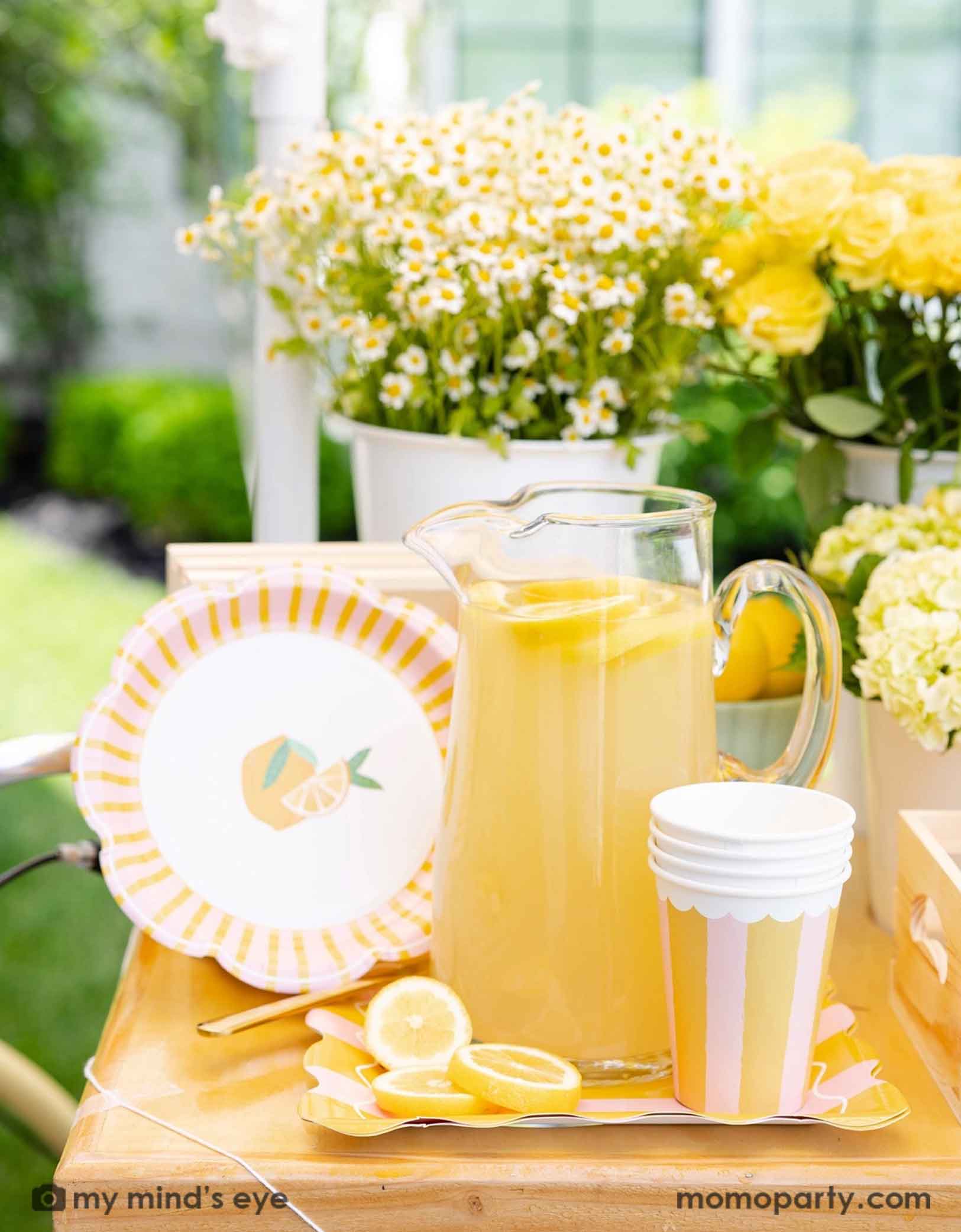A glass pitcher of lemonade with lemons and Momo Party's Yellow & Pink Striped Party Cups and pink and yellow striped side plates next to it. With bouquets of flowers in yellow and white on a wooden dessert cart with lemons in the background. A beautiful dessert cart for a summer-y lemon themed celebration, be it a kid's birthday party, a baby shower, or a bridal shower.  