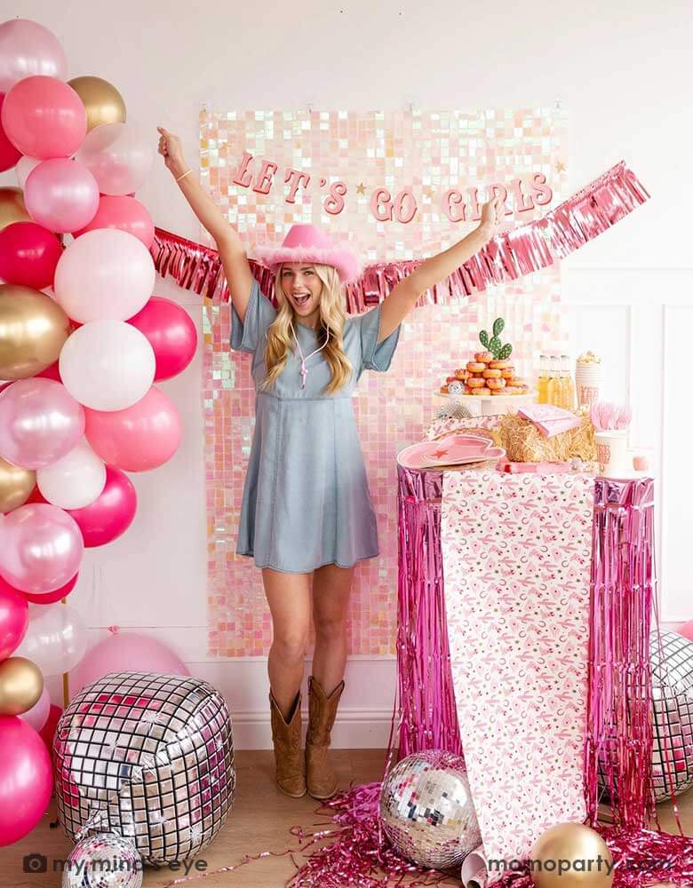 A young Caucasian lady in the Western inspired outfits and a pink cowgirl hat in a disco cowgirl bachelorette party decorated with cowgirl themed party supplies, decorations and balloons from Momo Party. She's raising her both hands by a table decorated with cowgirl party supplies including plates, napkins and a paper table runner. There's a donut cake, beverages and various treats on the table. Behind them is a iridescent wall adorned with "Let's Go Girls" pink foil fringe party banner from Momo Party.