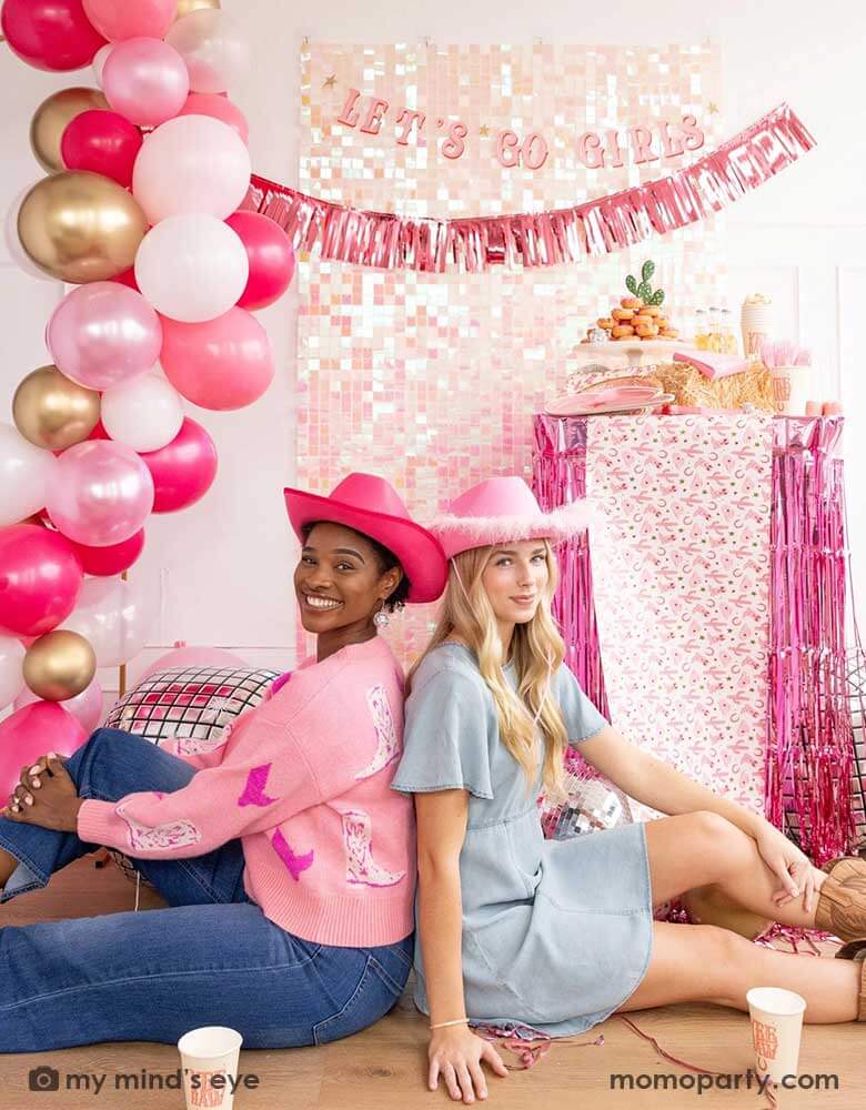 Two young ladies in Western inspired outfits and pink cowgirl hats in a disco cowgirl bachelorette party decorated with cowgirl themed party supplies, decorations and balloons from Momo Party. They're sitting back to back on the floor by a table decorated with cowgirl party supplies including plates, napkins and a paper table runner. There's a donut cake, beverages and various treats on the table. Behind them is a iridescent wall adorned with "Let's Go Girls" fringe party banner from Momo Party.