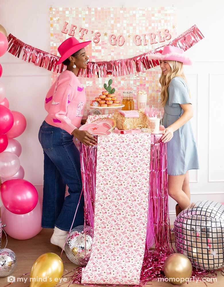 Two young ladies in their Western rodeo cowgirl inspired outfits and pink cowgirl hats in a disco cowgirl themed bachelorette party decorated with cowgirl themed party supplies, decorations and balloons from Momo Party. They're facing each other by a table decorated with cowgirl party supplies including plates, napkins and a paper table runner. There's a donut cake, beverages and various treats on the table. Behind them is a iridescent wall adorned with "Let's Go Girls" fringe party banner from Momo Party.
