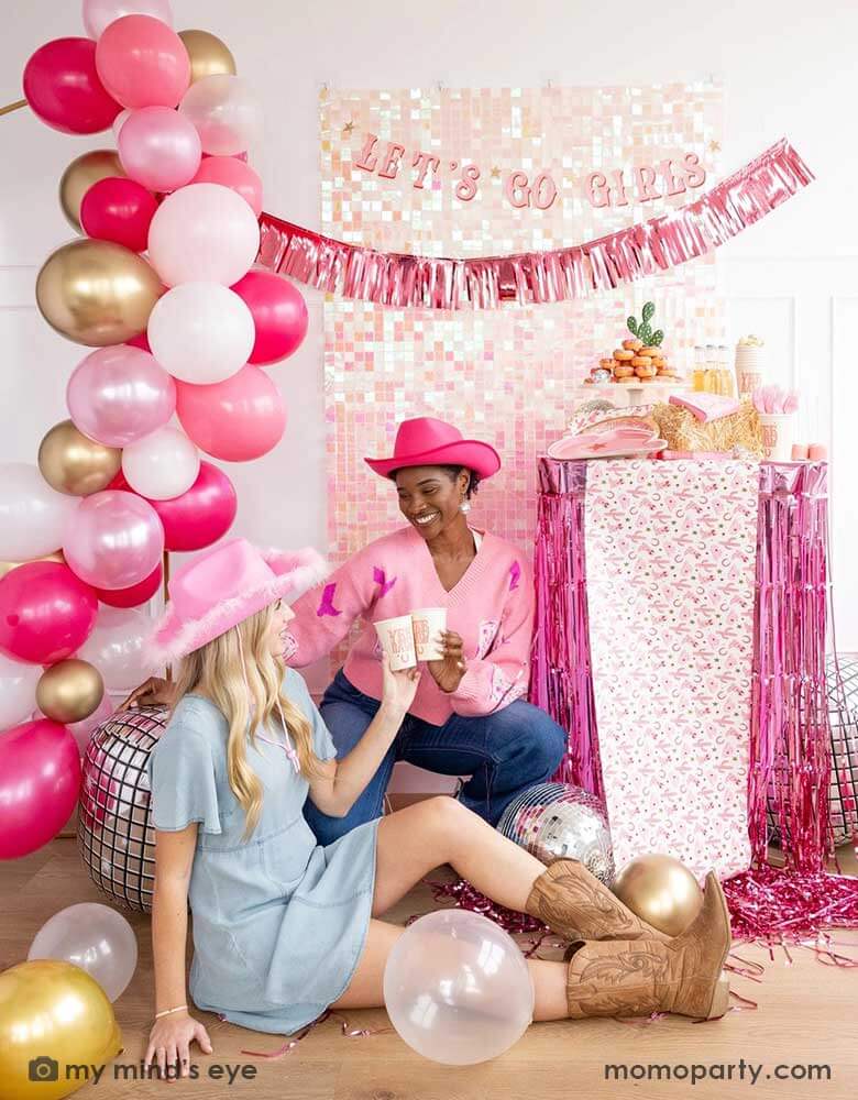 Two ladies with Western inspired outfits in a disco cowgirl bachelorette party decorated with cowgirl themed party supplies, decorations and balloons from Momo Party. They're raising their 12oz YEEHAW party cups by My Mind's Eye enjoying themselves in this festive celebration with donut cakes, beverages and various treats on the table decorated with cowgirl pattern table runner and pink foil fringe next to them. In the background there's a iridescent wall decorated with Let's Go Girls foil fringe banner.