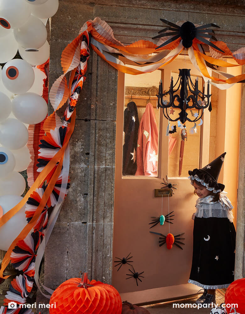 A spooky house with fainted lighting decorated with Momo Party's Halloween paper decorations including an eyeball balloon garland, honeycomb spiders, orange and pink crepe streamer garland, and a black chandelier. In front of the house there's a girl dressed up as a witch, she's looking at the spider decorations in a playful way.