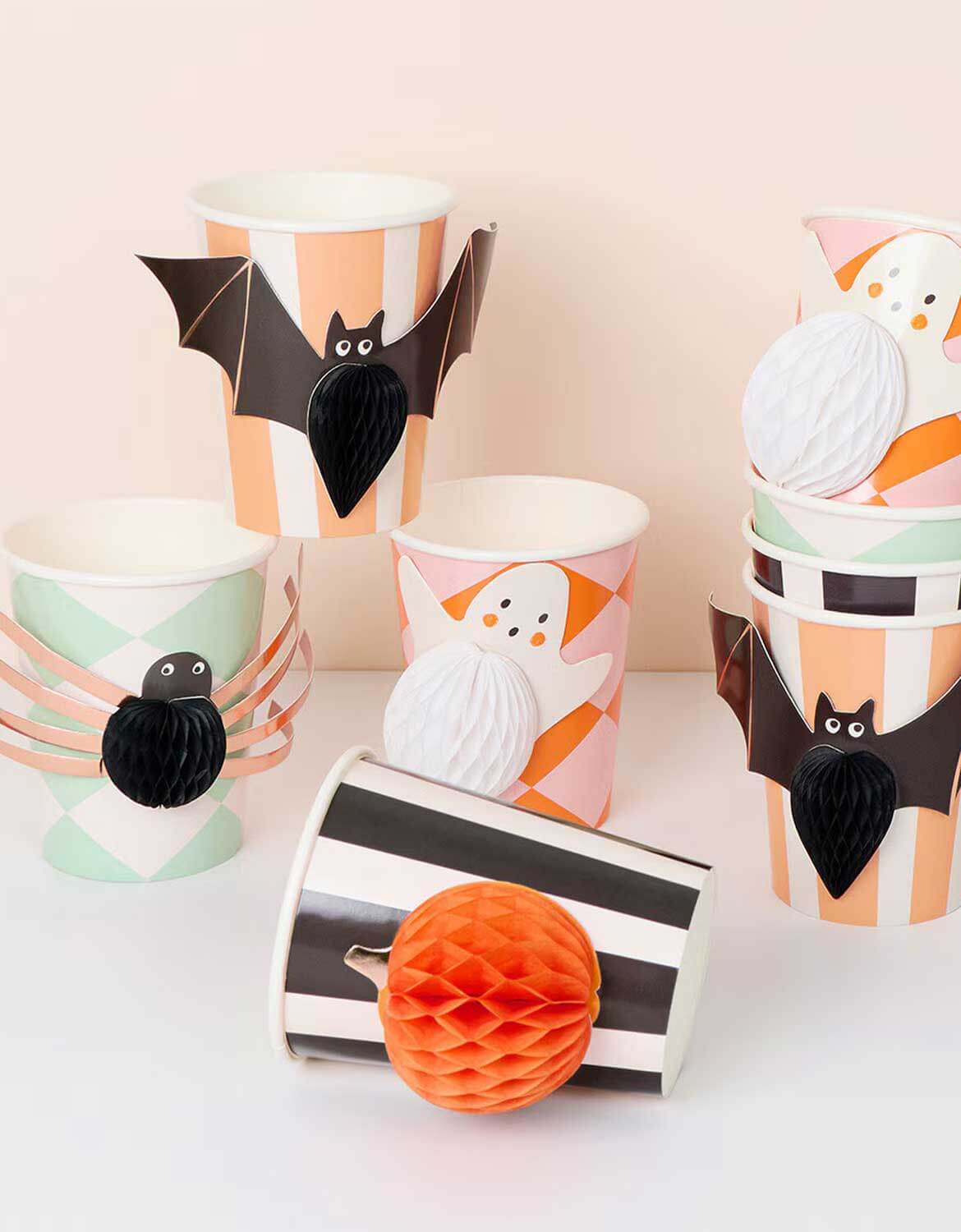 Momo Party's set of Halloween-themed honeycomb paper party cups decorated with bat, spider, and ghost graphics, styled with orange honeycomb paper balls on a soft pastel background.
