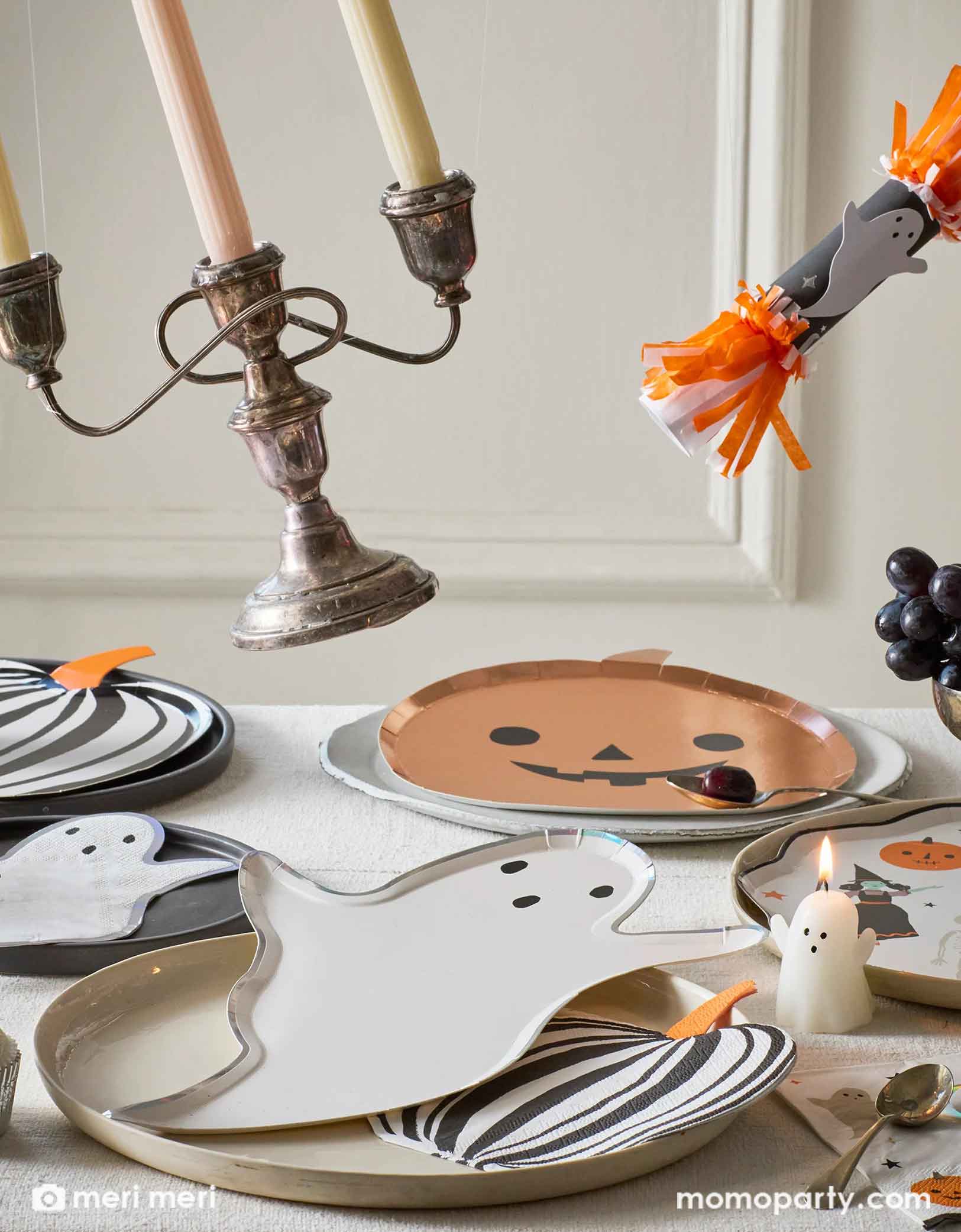 A Halloween-themed table setting with an array of Momo Party's Halloween party supplies including ghost and pumpkin shaped plates and matching napkins, black striped pumpkin plates and napkins by Meri Meri. With ghost candles, Halloween ghost party crackers and decorative items flying off on the table, this creates a fun scene for a kid's friendly Halloween celebration in this spooky season!