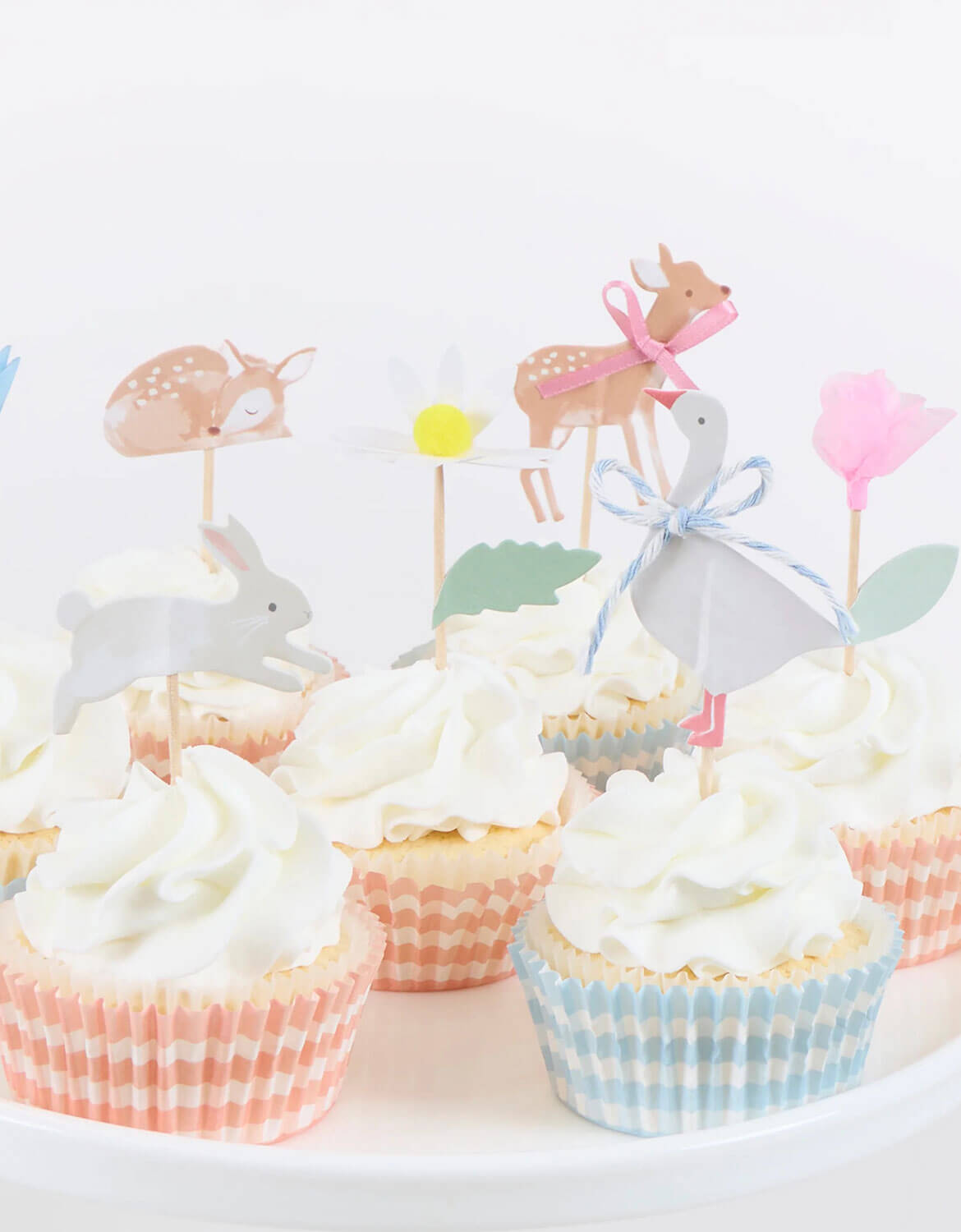 Cupcakes topped with Momo Party's set of little animals cupcake toppers of fawns, a rabbit, a goose, and flowers on sticks by Meri Meri against a white background.on a white background.